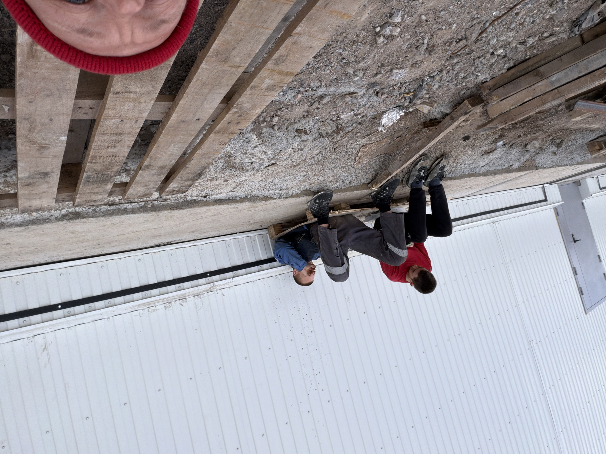An outdoor scene captures three individuals in what appears to be a work or storage yard in Chaikovsky, Russia. The image is taken from a low, close perspective, with the upper portion dominated by the forehead and eyes of a person wearing a red knitted hat, likely the photographer.
Below this, several wooden pallets are scattered and loosely stacked on a coarse ground made of gravel, dirt, and small stones. Two other individuals are seated or crouching among these pallets. One person, wearing a blue jacket and grey work pants, is seated on a pallet. The other individual, in a red long-sleeved shirt and black pants, is also near the pallets, with their back partially turned towards the camera. Both appear to be resting or observing.
The background features a large, white corrugated metal building with vertical panels and visible fasteners. A plain grey door with a black handle is visible on the far right side of the building. The overall lighting is soft and even, suggesting an overcast day. There is no discernible text visible in the image.