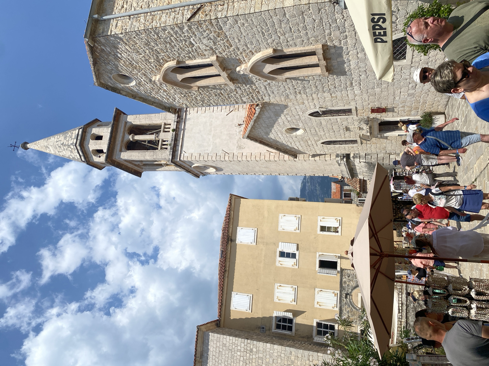 The image captures a street scene in Budva, Montenegro. The foreground features a bustling marketplace with vendors displaying goods under large umbrellas. Several people are browsing the merchandise. To the left, a tan-colored building with white-shuttered windows rises above a stone structure. Ascending into the sky, a stone bell tower with a pointed spire stands prominently against a backdrop of blue sky and scattered clouds. Another stone building adjoins the tower, featuring arched windows and a "Pepsi" branded sunshade. People are walking along the street near the stone buildings. The image appears to be taken at ground level, looking upwards towards the architecture and sky.