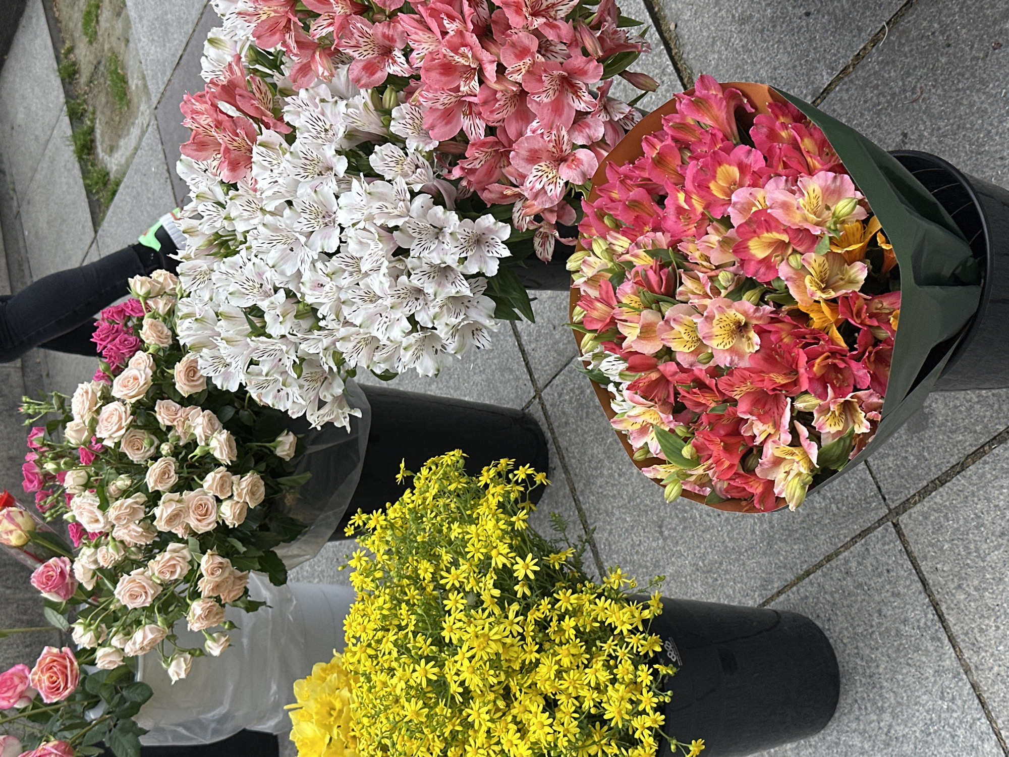 A variety of cut flowers in black plastic buckets are on a sidewalk. The flowers include pink and white alstroemeria, roses, and small yellow flowers. The sidewalk is paved with rectangular stone blocks. The photo was taken in Tbilisi, Georgia. A person wearing black leggings is partially visible on the left side of the image.