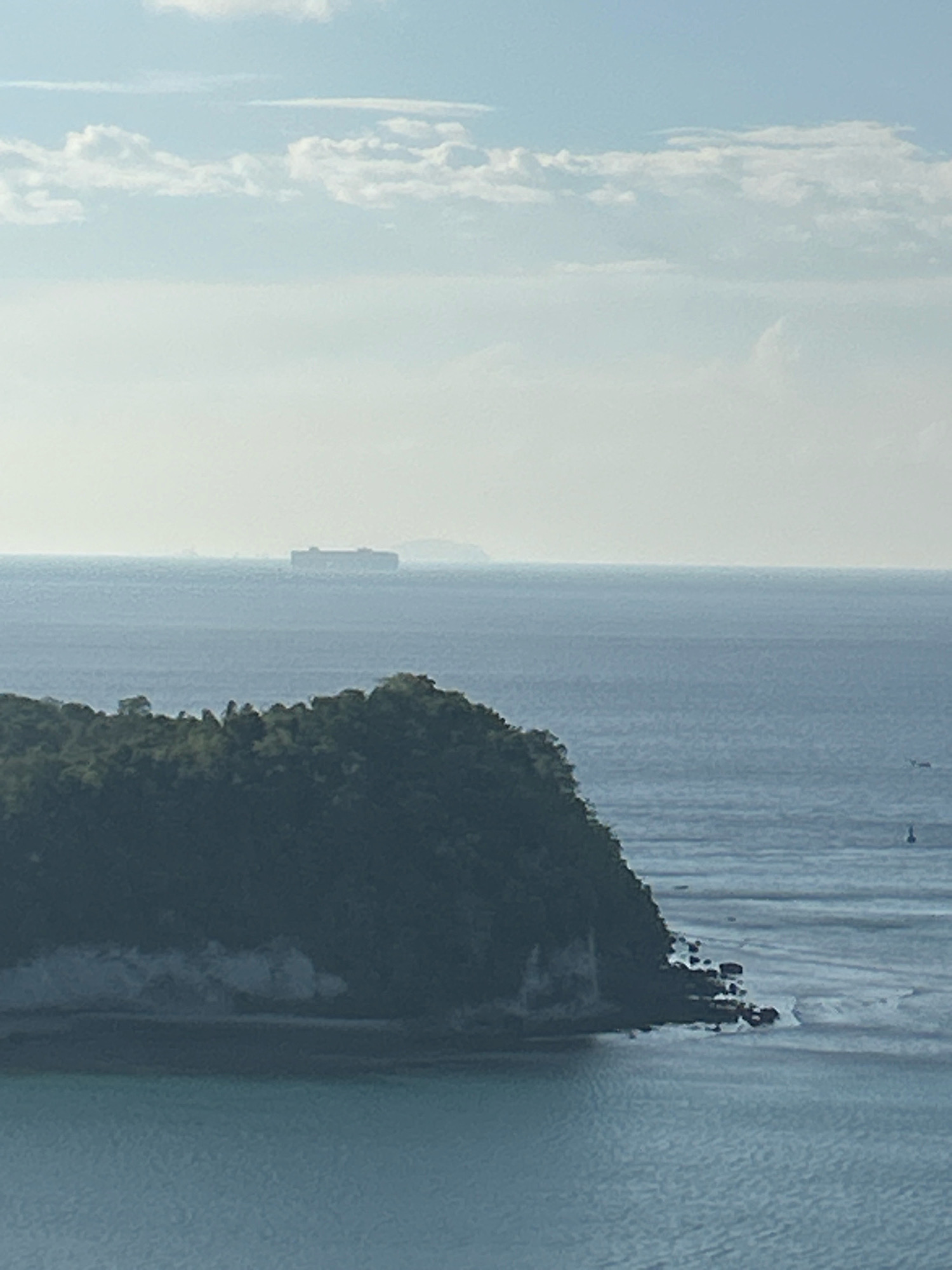 The image depicts a vast expanse of the ocean under a hazy sky with scattered clouds. In the foreground, a lush, green, tree-covered island juts out into the water, its rocky shoreline marked by small waves. The island appears to be part of the city of Sabang, Indonesia, a location known for its natural beauty and maritime environment. Further out at sea, a large, rectangular cargo ship is visible on the horizon, a tiny speck against the immense blue backdrop. The overall impression is one of serene tranquility, with the immensity of nature dominating the scene.