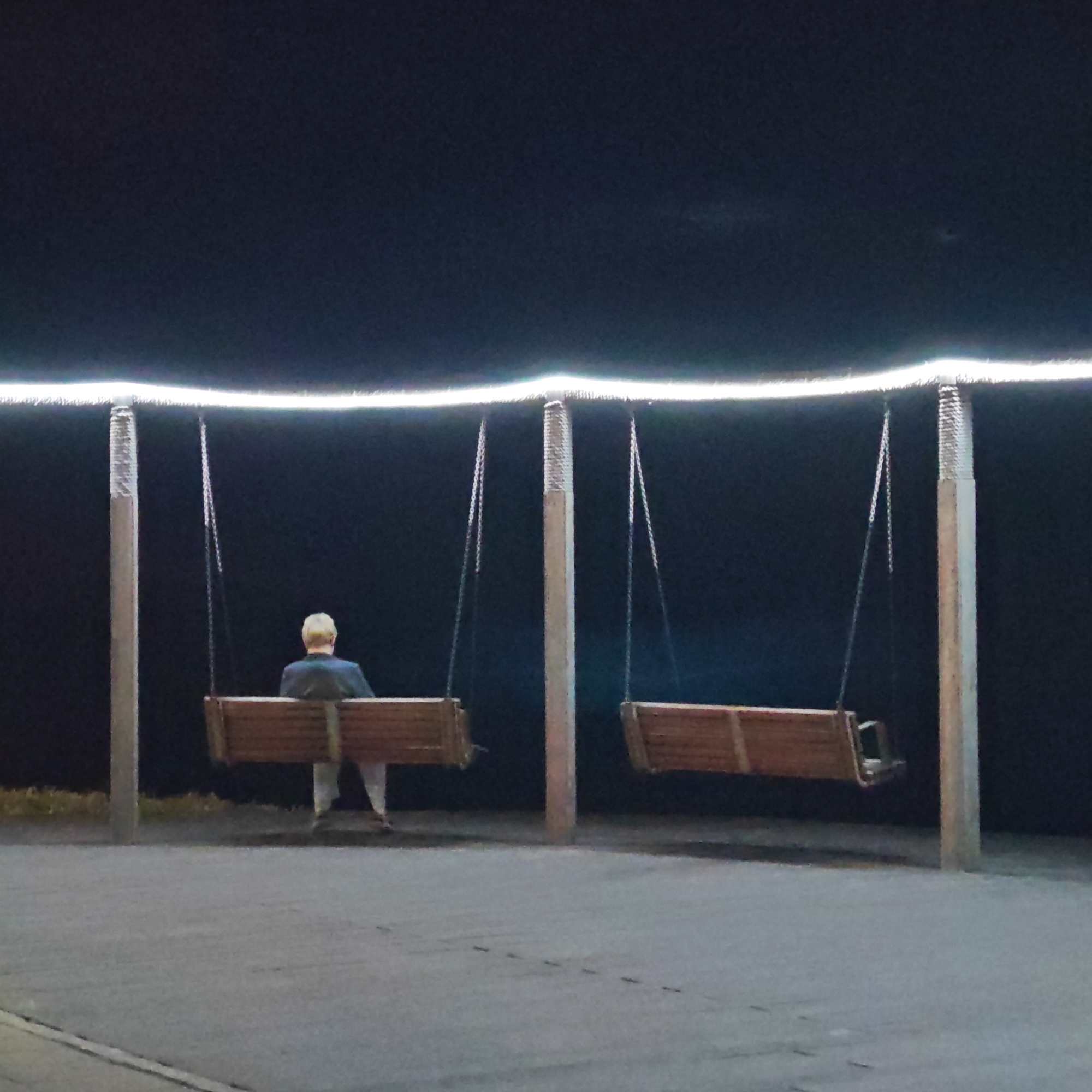 The image depicts an outdoor nighttime scene in городское поселение Щелкино, Ukraine. The main subjects are a person and two wooden swings, illuminated by an overhead light strip.
A single individual, seen from behind, with light-colored hair and wearing a dark top, sits on the left swing. Their posture suggests they are at rest, not actively swinging. The second swing to the right is empty. Both swings are suspended by chains from a structure composed of three vertical poles. These poles appear to have a patterned texture towards their tops.
Above the swings, a bright, continuous strip of white light forms a gentle, illuminated arch against the very dark, almost black, night sky. This artificial light source provides the primary illumination for the scene, highlighting the swings and the person. The ground beneath the swings is a light-colored paved surface. Faint traces of grass or low vegetation are visible in the dark background directly behind the swings.
The scene conveys a sense of quiet solitude or contemplation, with the lone figure under the artificial light in the dark surroundings. There is no discernible text visible in the image.