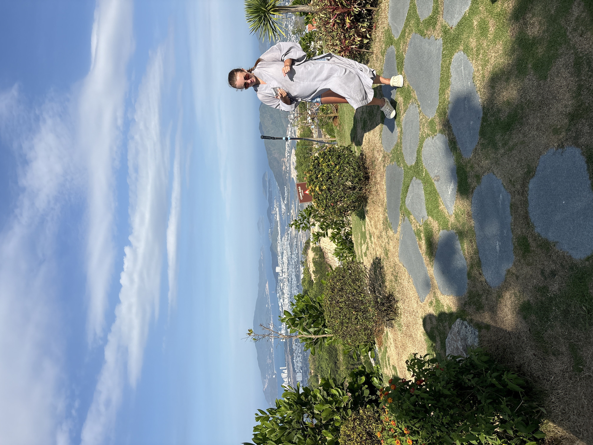 The image shows a woman standing on a stone pathway in Tay Nha Trang Ward, Vietnam. She is wearing a grey robe over denim shorts and white clogs, holding a phone in her hand. The pathway is surrounded by grass and greenery. Behind her is a panoramic view of the city, mountains, and the sea. The sky is blue with scattered clouds.