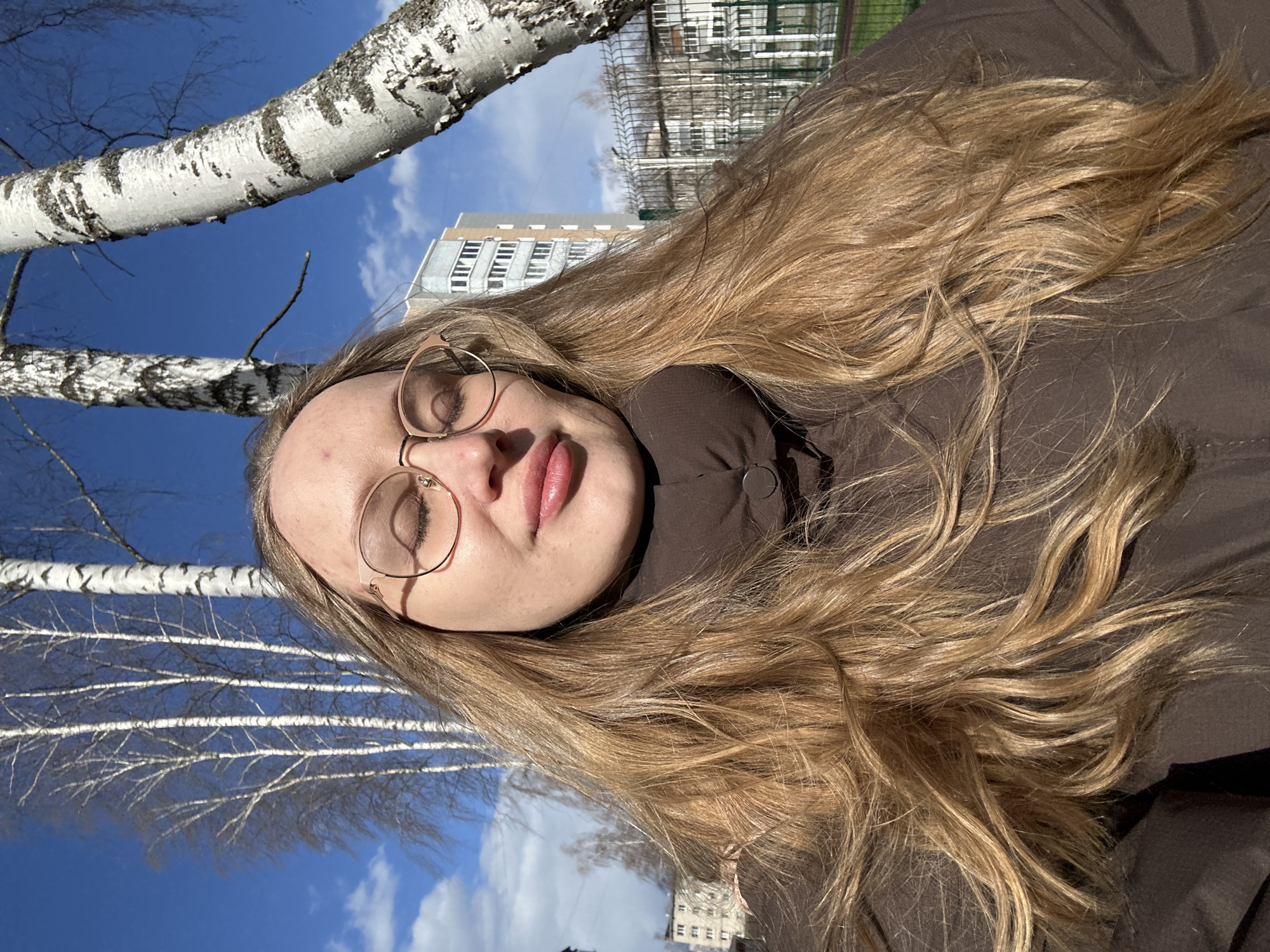 An outdoor portrait shot in Chaikovsky, Russia, features a young woman with long, flowing blonde hair, captured from a slightly upward angle and rotated 90 degrees counter-clockwise. The woman's eyes are closed, and her lips are slightly parted, conveying a serene and relaxed expression. She is wearing thin, round-rimmed glasses and a brown, possibly padded jacket.
The background shows a clear, vibrant blue sky with scattered white clouds. Several birch trees with their characteristic white and dark bark and bare branches are prominently visible, suggesting the season is late winter or early spring. In the distance, a multi-story building, likely a residential apartment complex, is visible, along with a green fence or netting. The bright sunlight creates strong highlights on her hair and illuminates the scene, indicating it is daytime. No discernible text is visible in the image.