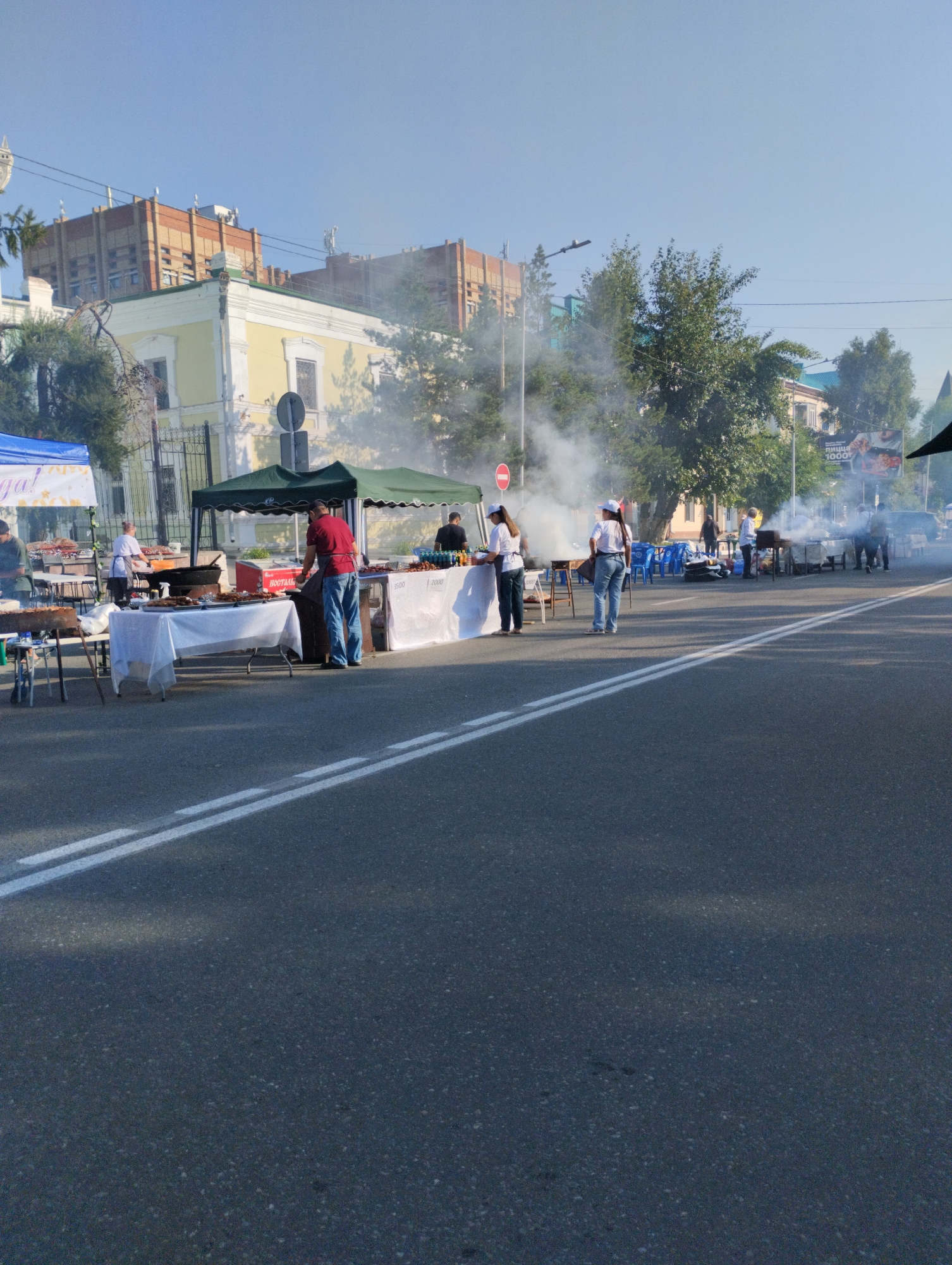 The image shows an outdoor food market scene on a street in Kostanay, Kazakhstan. Several tents are set up, providing shade for vendors and displaying food items, primarily grilled meats. Smoke is rising from grills, indicating that food is being prepared. People are seen walking around and interacting with vendors. In the background, there are buildings characteristic of urban architecture, including some older, ornate structures and more modern ones. The street is paved and has white lane markings. The sky is clear and blue, suggesting a sunny day. The overall impression is of a lively street market event.
