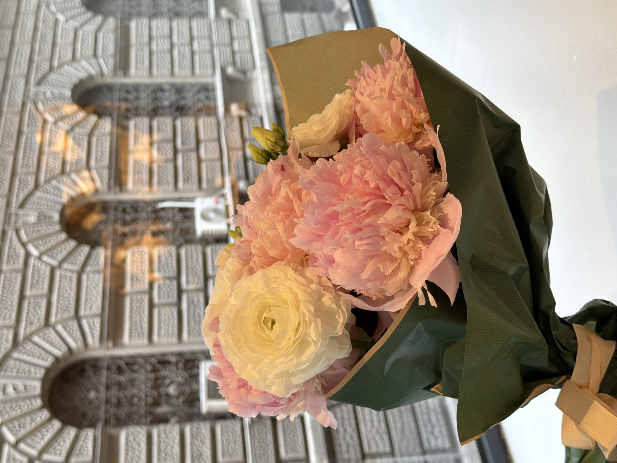 A bouquet of flowers is the central subject of the image. The bouquet features large, pink flowers, possibly peonies, and a single, prominent white ranunculus. The flowers are wrapped in green and brown paper, tied with a light-colored ribbon.
The bouquet is held up against a window, which reflects a view of the exterior. The reflection shows a gray, ornate building facade, likely made of stone or concrete. The architectural design includes repeating arched elements.
Without additional visual cues, specific identification of the exact location is not possible but the style suggests a historical building in the city of Belgrade, Serbia.
