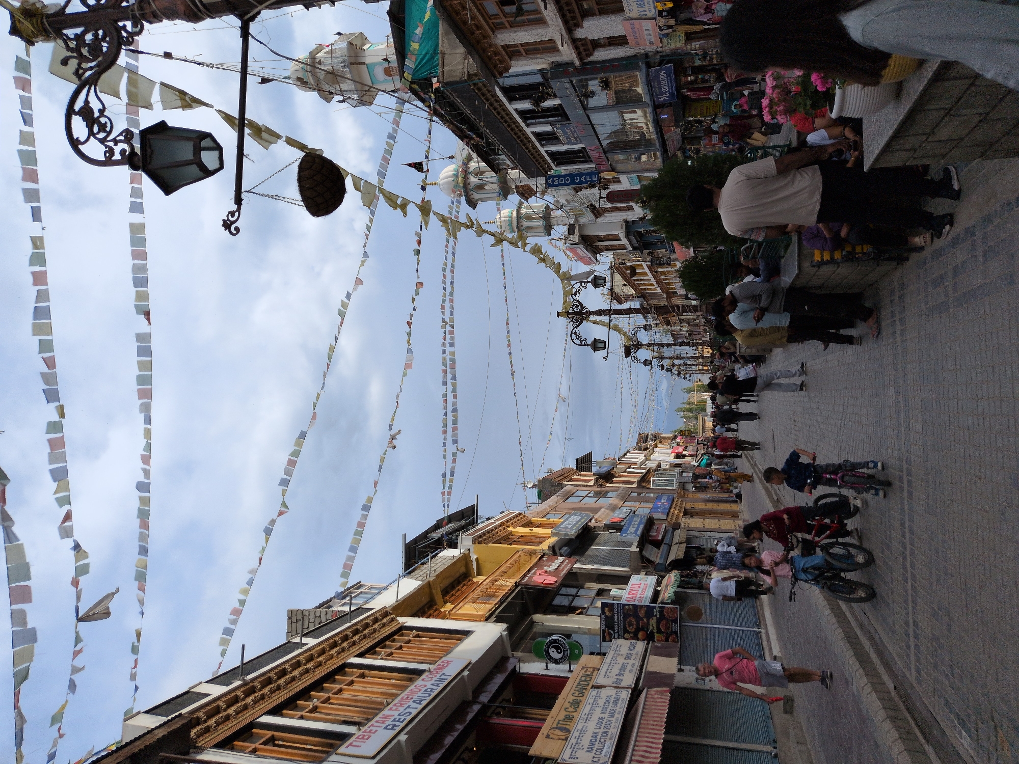 The image depicts a bustling street scene in Leh, India, likely a main thoroughfare lined with shops and businesses. Prayer flags are strung across the sky, creating a festive and cultural atmosphere. The architecture is a mix of traditional and more modern styles, with multi-story buildings housing various shops. The street is paved and appears to be a pedestrian-friendly area, with several people walking and others engaged in activities.
In the foreground, three children are near bicycles, with two girls on bicycles and a boy standing next to a pink bicycle. A man in a pink t-shirt and grey shorts stands to the left of the children, facing the street. Further down the street, numerous people are walking, some singly and others in groups, browsing shops or moving along. A prominent street lamp with decorative metalwork is visible on the left side of the image. The backdrop includes buildings with visible signage, some of which are in English, indicating commercial activity. The overall impression is one of a vibrant and active urban environment in Leh, characterized by its unique cultural elements like the prayer flags.