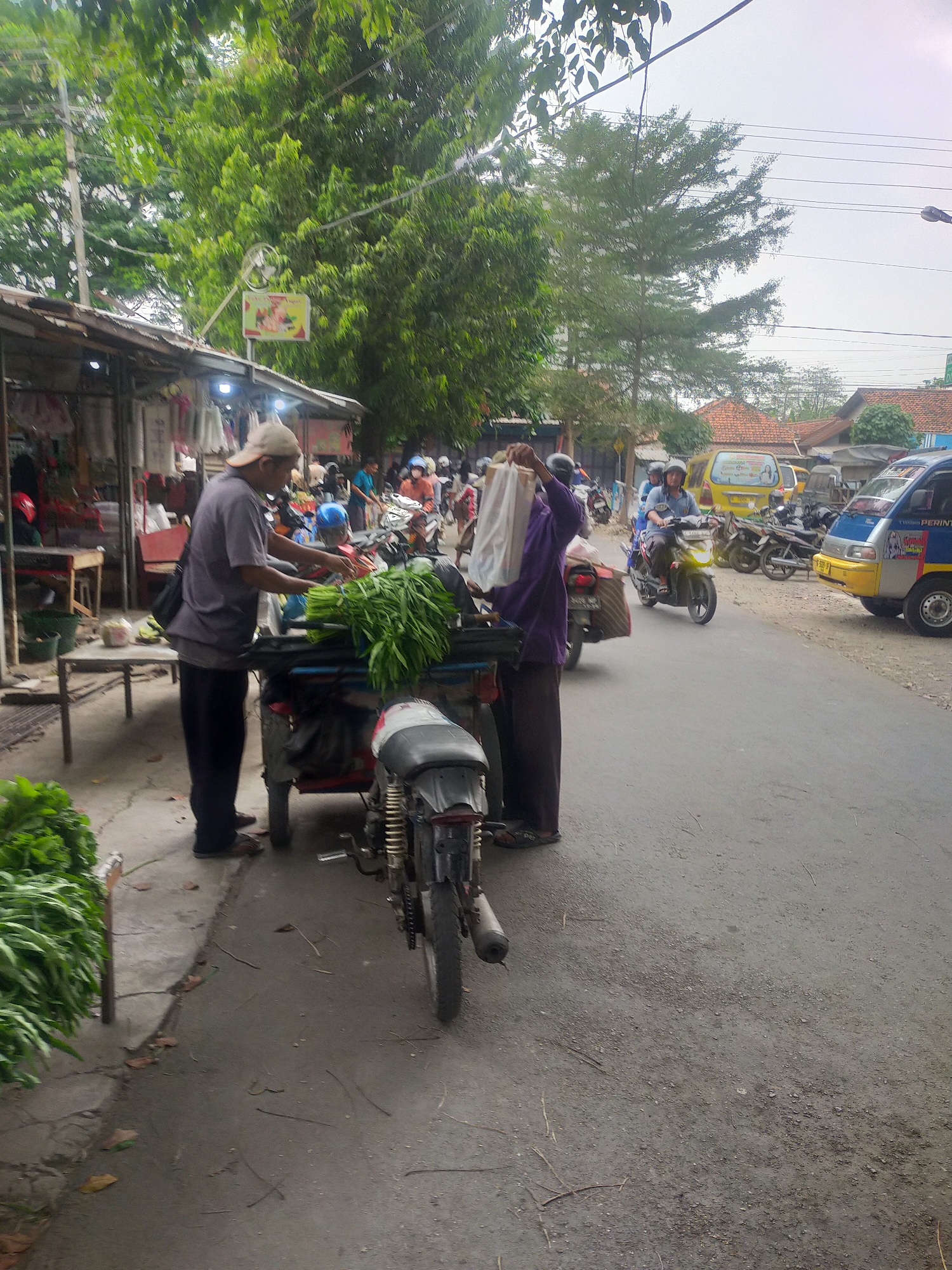 In Slawi, Indonesia, on an overcast day, a man wearing a baseball cap and dark pants stands beside a cart loaded with green vegetables, seemingly attending to his produce. He is near a roadside stall with an awning, where goods are displayed. A woman in a purple shirt and dark pants walks away from the cart, holding a white plastic bag. Other individuals and motorcycles are visible on the street, indicating a bustling market or roadside scene. Vehicles, including a yellow van and a blue and yellow truck, are parked or driving on the road. The environment is characterized by trees, overhead wires, and buildings with tiled roofs.