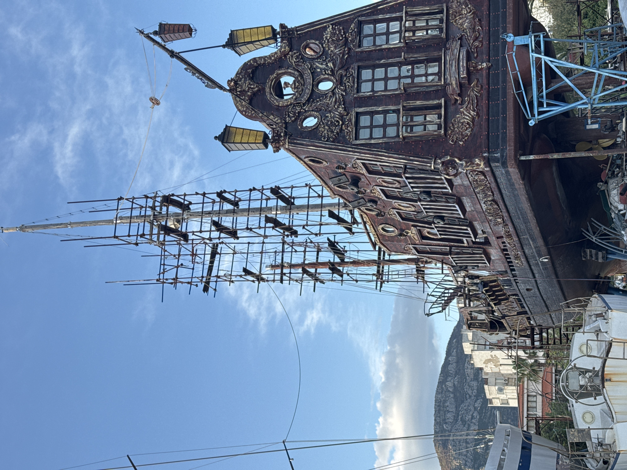The image shows the stern and rigging of a large, ornate, brown, partially-constructed ship. The ship is docked in the city of Bar, Montenegro, close to land and other boats. The stern has multiple levels with windows, and ornate carvings. The rigging is a framework of poles and cross-beams. The sky is blue with some white clouds. In the background are buildings and a mountain. There is blue scaffolding attached to the side of the ship.