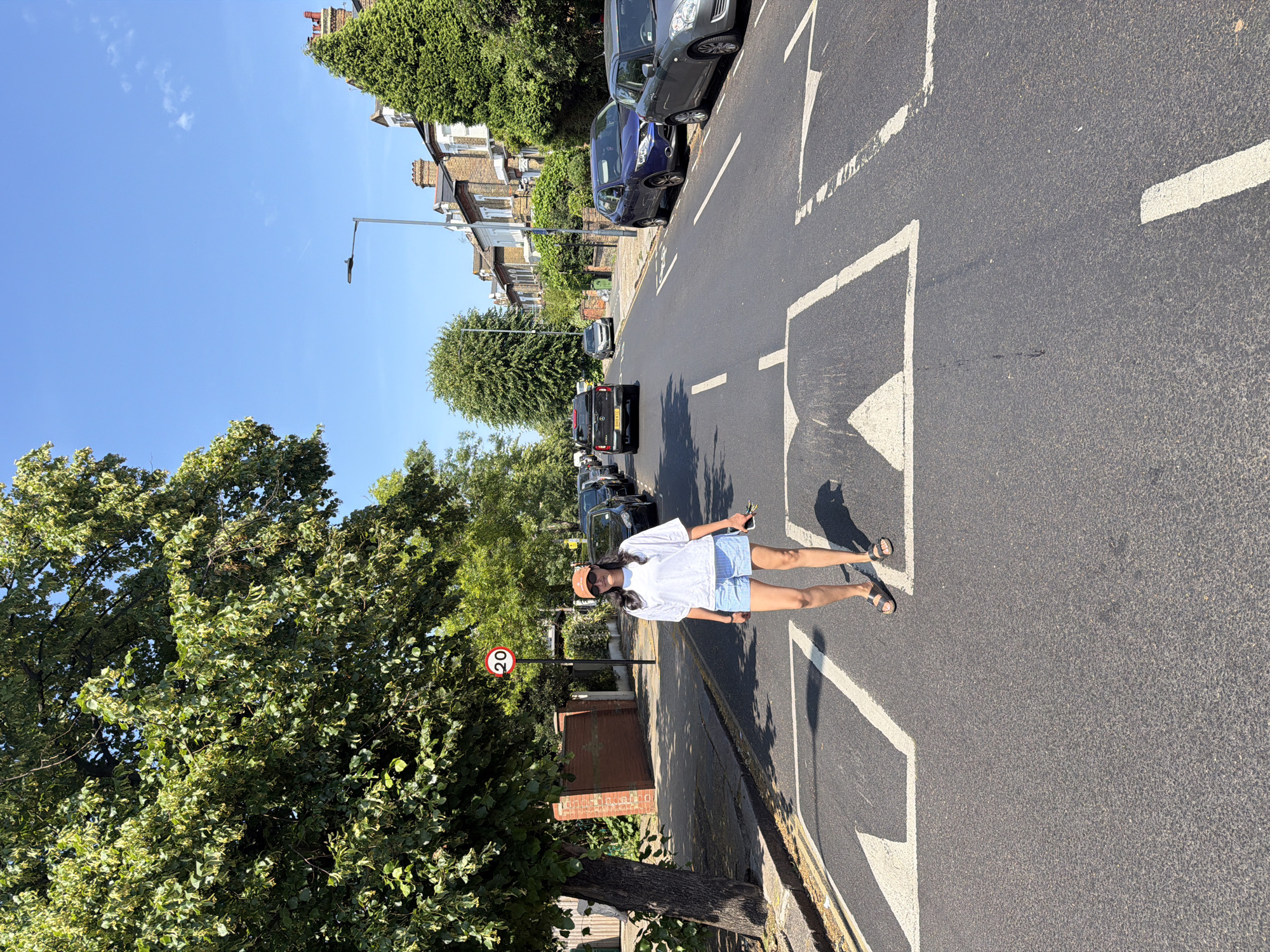 A full-body shot captures a young woman standing on a residential street. She is wearing a white t-shirt, blue striped shorts, sunglasses, and a tan hat. She holds a phone in her right hand and appears to be looking forward. Her left foot is slightly forward, and she is wearing sandals.
The setting is a street in London, United Kingdom, indicated by the architecture of the houses and the presence of a "20" speed limit sign. The houses are made of brick and have traditional pitched roofs, with trees lining the street. The road has clear white markings, including a pedestrian crossing. Several cars are parked along the street and some are driving on the road, including a black van.
The sun is shining brightly, casting shadows on the road, suggesting it is daytime. The sky is a clear, bright blue, indicating fair weather. The overall mood of the image is casual and relaxed, with the woman enjoying a sunny day outdoors.