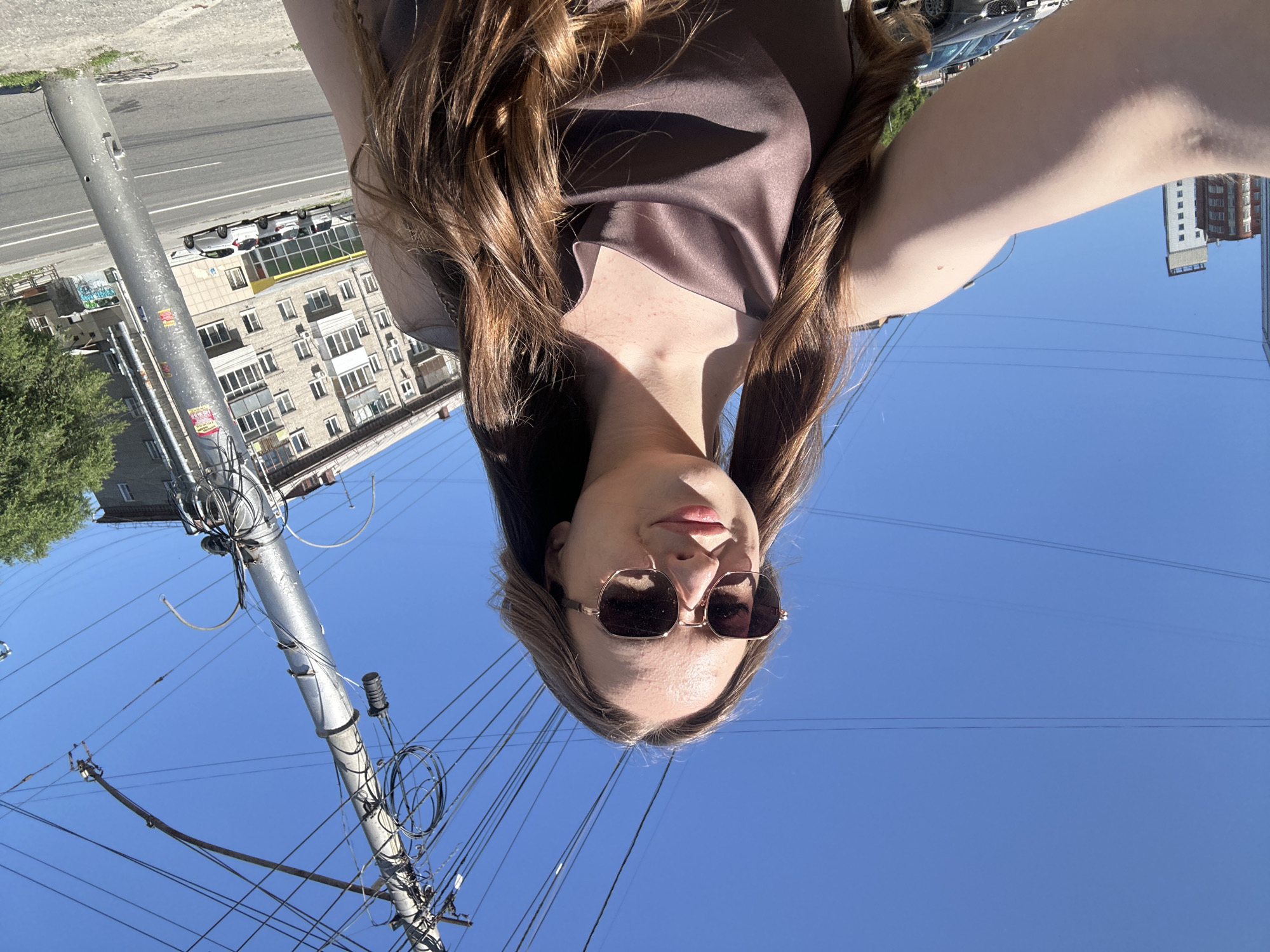 A young woman with long, wavy brown hair and sunglasses is taking a selfie outdoors on a sunny day. She is wearing a shimmering brown dress. She is standing on a sidewalk next to a street with cars parked on the side. In the background, there is a tall utility pole with many wires, a multi-story brick building, and a clear blue sky. The scene appears to be in an urban environment.