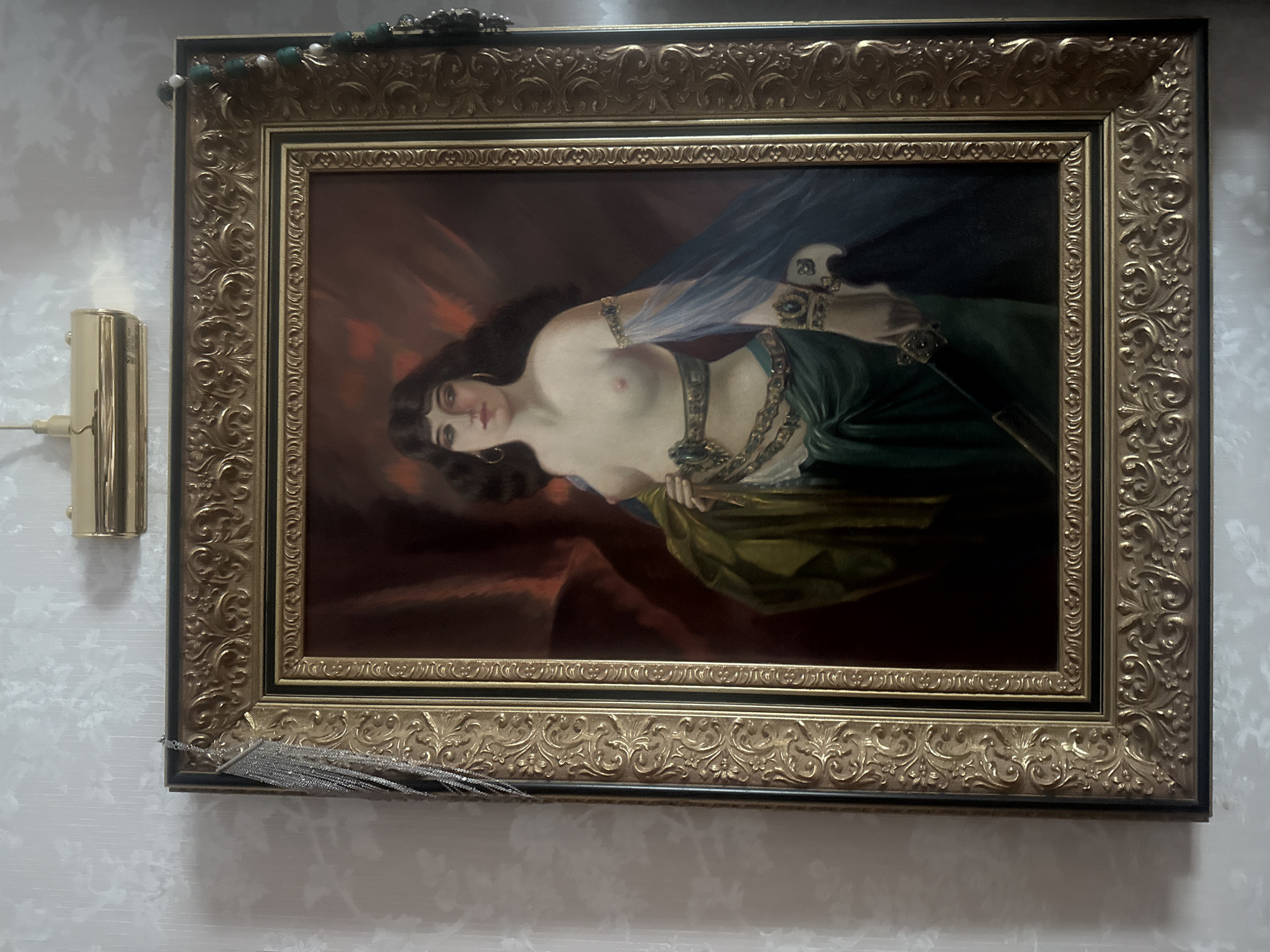 A framed painting hangs on a patterned wall. The painting depicts a reclining, semi-nude woman with dark hair, draped in fabrics and adorned with jewelry. The painting is housed in a gilded, ornate frame with a secondary black frame. A gold-colored light fixture is visible on the wall to the left of the painting. A decorative tassel hangs from the bottom of the frame. The location cannot be determined based on the visual information provided.