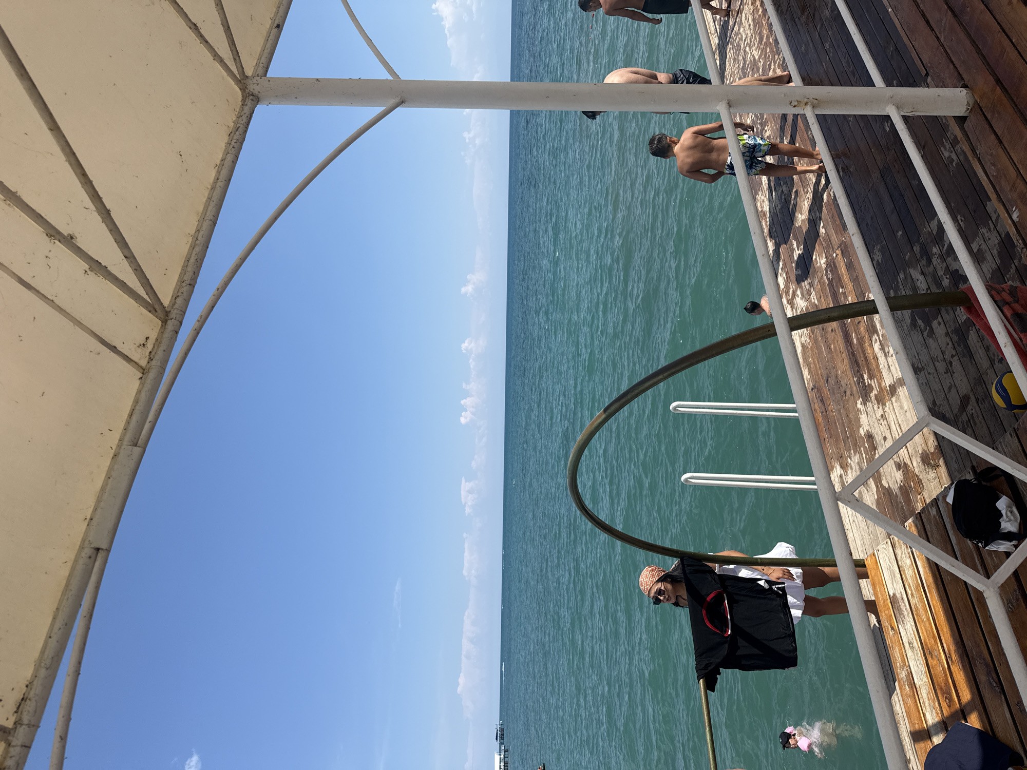 The image depicts a bright, sunny day on a wooden pier overlooking a large body of water, likely Lake Issyk-Kul, in Sary-Oy, Kyrgyzstan. The water is a vibrant teal color and appears calm with gentle ripples. In the foreground, a wooden pier is visible, with a white metal railing running along its edge. A woman wearing a headscarf and sunglasses stands near the railing, holding a black t-shirt. A young child in a pink swim cap is in the water to her left. Further down the pier, several shirtless boys and men are standing, some appearing to be preparing to enter the water. One boy is facing away from the camera, wearing patterned swim shorts. The sky above is a clear blue with a few scattered white clouds. A white canopy structure is partially visible in the upper portion of the image, providing shade. The overall scene suggests a recreational activity, likely swimming or sunbathing, at a lakeside resort or public beach.