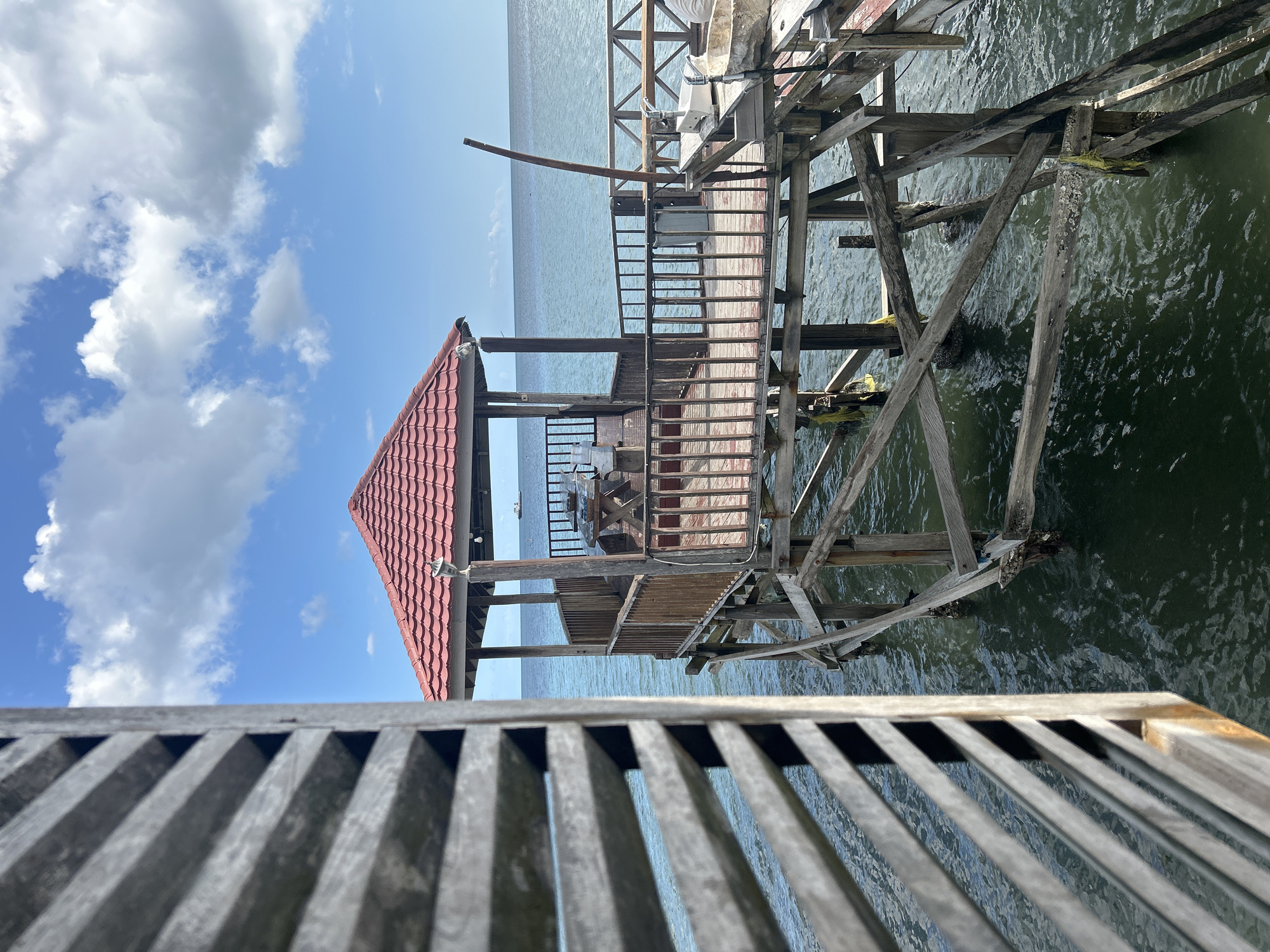 A wooden gazebo with a red tiled roof stands on stilts over the ocean. The gazebo is accessible via a wooden walkway and features wooden railings, a table, and benches. A ship is visible on the horizon. The scene is framed by wooden shutters on the left side of the image. The location is not specified in the city of A R A, Indonesia.