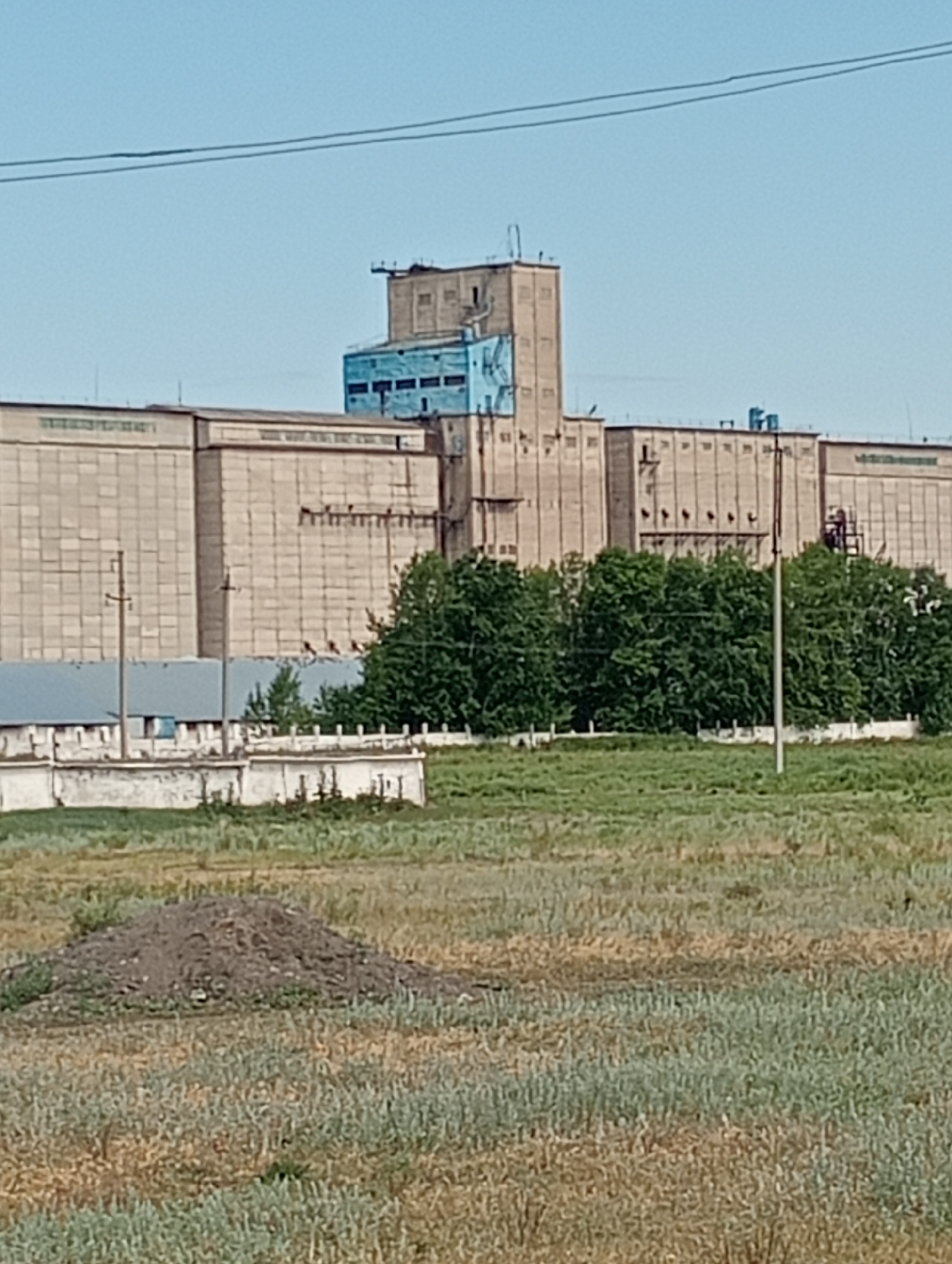 An industrial complex with a towering silo structure dominates the background of this outdoor, daytime scene. The silo, painted in sections of tan and bright blue, rises above a long, low-slung industrial building with a textured, tan facade. This main building appears to be an industrial facility, possibly a factory or warehouse, with a series of evenly spaced windows or vents along its upper sections. A line of green trees stands in front of the industrial building, creating a natural barrier between the structure and the foreground. The foreground consists of a grassy field with a dirt pile in the lower-left quadrant. The sky is a clear, pale blue with two thin, dark lines, possibly power lines, stretching horizontally across it. The overall environment suggests an industrial area on the outskirts of a town or city.