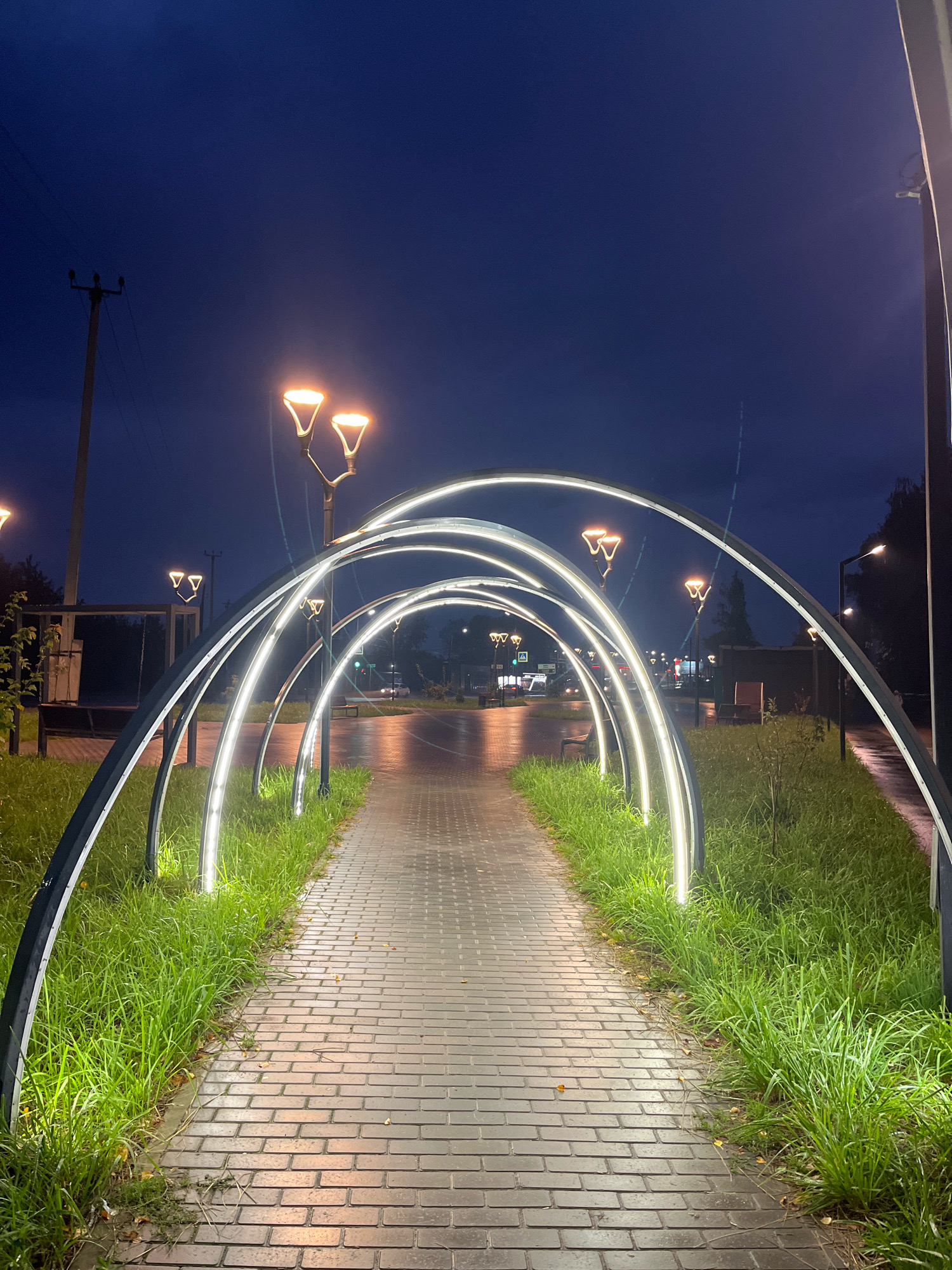 A pathway lined with illuminated arched structures stretches into the distance. The pathway is paved with rectangular bricks and is flanked by lush green grass. The arched structures, with white LED lighting, create a tunnel-like effect. In the background, streetlights with orange glow illuminate the scene. A utility pole with wires stands to the left of the pathway. Traffic lights and a few distant vehicles are visible on a wet road. The sky is a deep blue, indicating it is nighttime or twilight. The overall scene suggests an outdoor public space, possibly a park or a pedestrian walkway in the city of Chaikovsky, Russia.