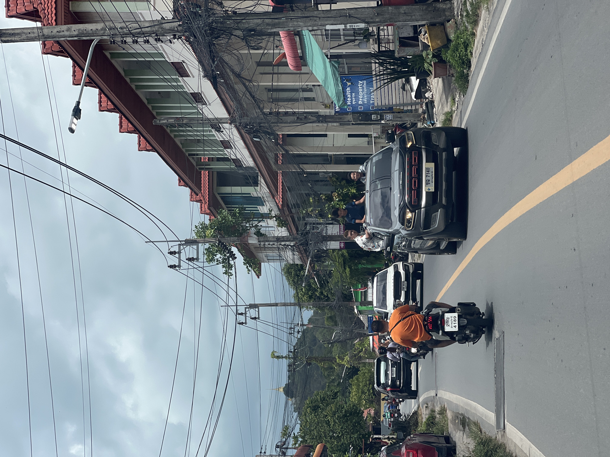 The image depicts a street scene in an urban area, likely in Thailand, based on the vehicle license plates and signage. There is a congested network of overhead power lines. A black Ford truck is prominent in the foreground, with people standing nearby. A motorcyclist wearing an orange shirt is positioned in the center of the frame, heading in the same direction as the truck. A row of parked or slow-moving cars lines the side of the road. Buildings with red-tiled roofs are visible along the street. A billboard with text in Thai and English about 'Health, Property, Travel' is attached to one of the buildings. In the background, a hill with trees and a temple or structure is visible. The sky is overcast.