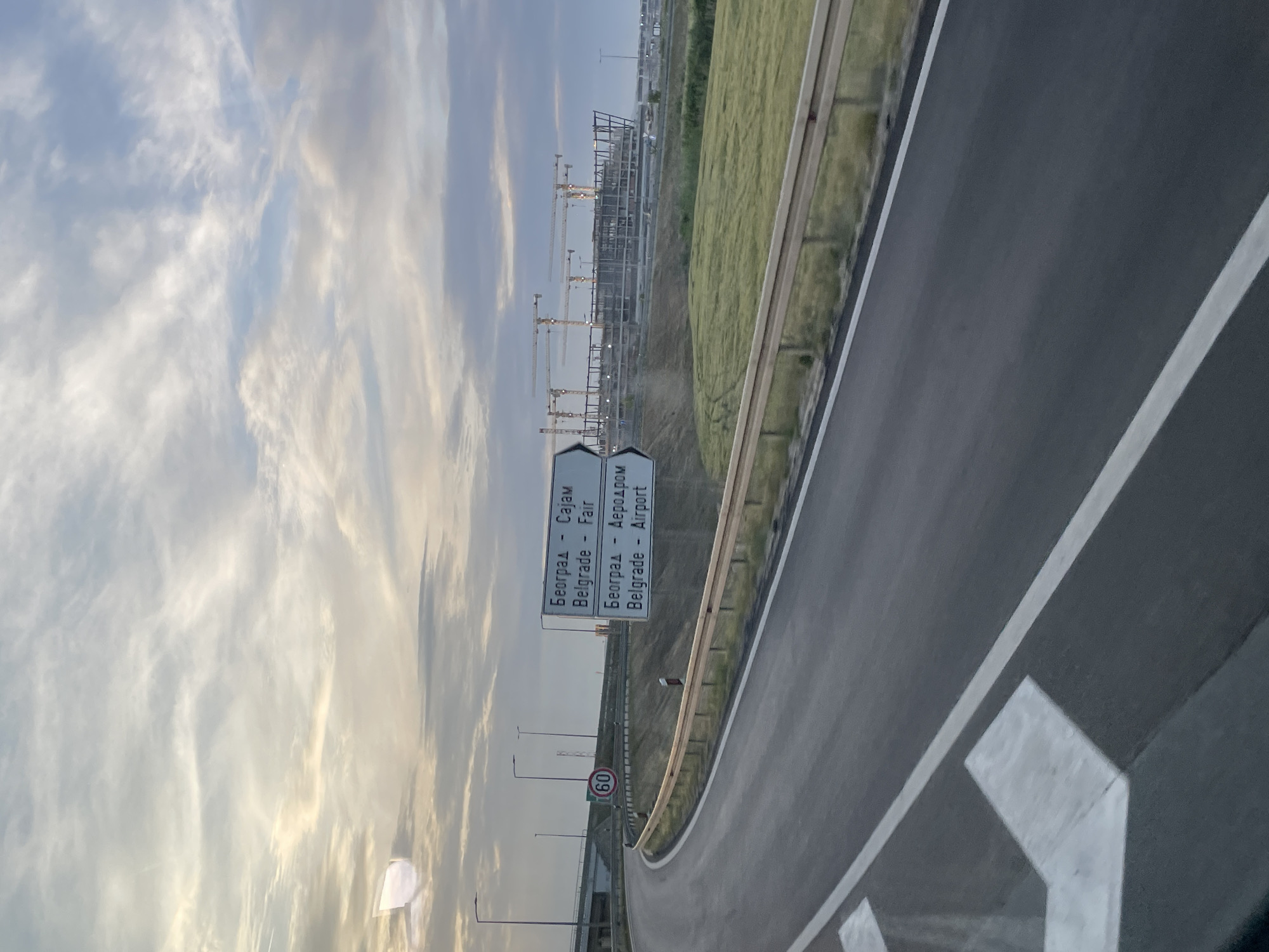 The image shows a view from a car window on a highway leading towards Belgrade, Serbia. Road signs indicate directions to Belgrade Fair and Belgrade Airport. Construction with numerous cranes is visible in the distance. The sky is filled with clouds. A speed limit sign shows 60. The location is likely in Surcin Urban Municipality, Serbia, based on the road signs pointing to Belgrade Airport, which is located in this municipality.
