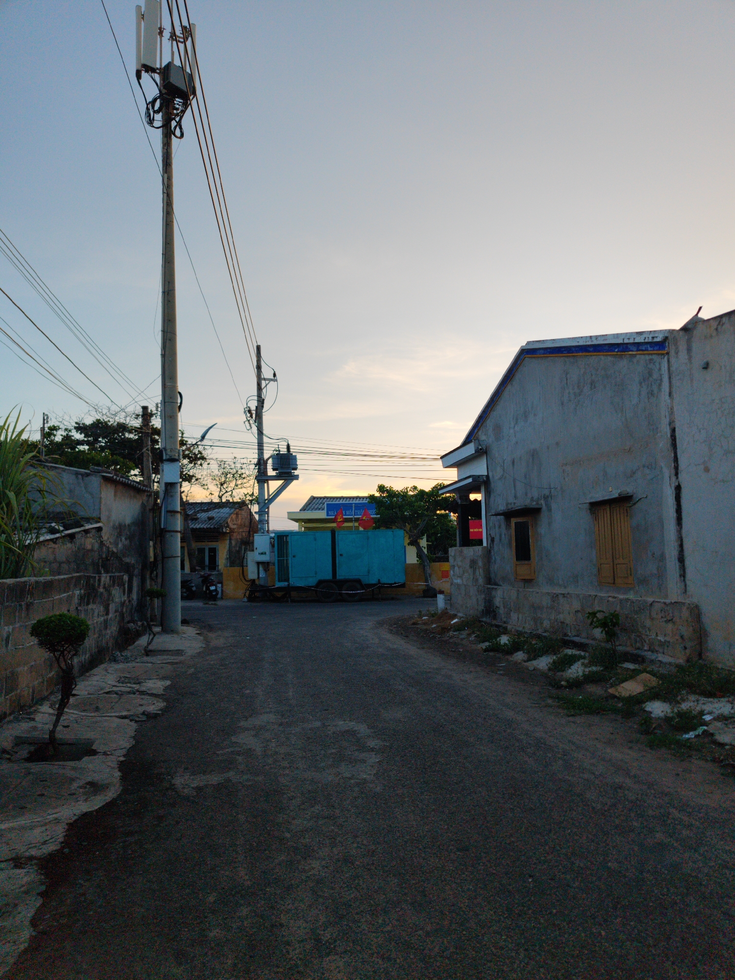 A narrow, asphalt road curves between buildings in Phú Quý Special Administrative Region, Vietnam. A tall utility pole with cellular antennas dominates the left foreground, accompanied by numerous power lines overhead. A low stone wall runs along the left side of the road, punctuated by small trees. On the right, a weathered concrete building with two shuttered windows stands close to the road. In the distance, a blue portable generator trailer with Vietnamese flags is visible. The sky is light, suggesting either dawn or dusk.