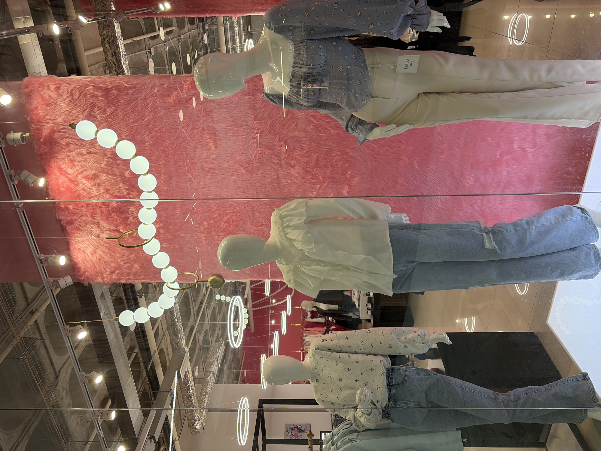 The image shows a retail display window, likely located in a shopping mall or commercial district. Three mannequins are visible, each wearing a different outfit. The top mannequin is dressed in a light blue floral-patterned top and white pants. The middle mannequin is wearing a white blouse and ripped blue jeans. The bottom mannequin is wearing a white and blue floral top and blue jeans. The backdrop of the display is a bright pink, shaggy material. Above, the ceiling of what appears to be a mall is visible, featuring a complex network of pipes, lighting fixtures, and structural supports. Additional clothing and mannequins can be seen in the background, through the glass window. The reflections visible on the glass window suggest the picture was taken inside a shopping mall. Based on the "Je Lo" tag on the white pants of the top mannequin, it is likely that the display is in Izhevsk, Russia.