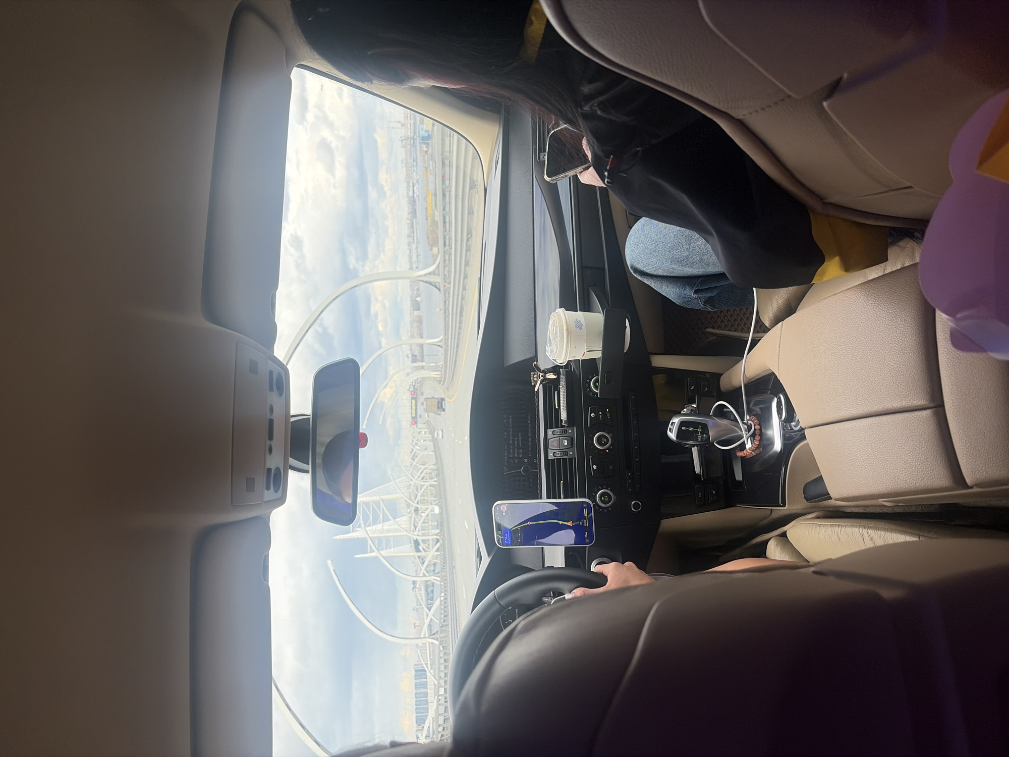 The photo was taken from inside a car traveling on a bridge in Saint Petersburg, Russia. A driver is visible at the steering wheel. The passenger is using a cell phone. The car's infotainment system and a navigation app on a phone mounted to the dashboard are visible. Through the windshield, the distinctive architecture of the Western High-Speed Diameter (WHSD) highway, with its complex interchange and cable-stayed bridge, is visible against a sky with patchy clouds.