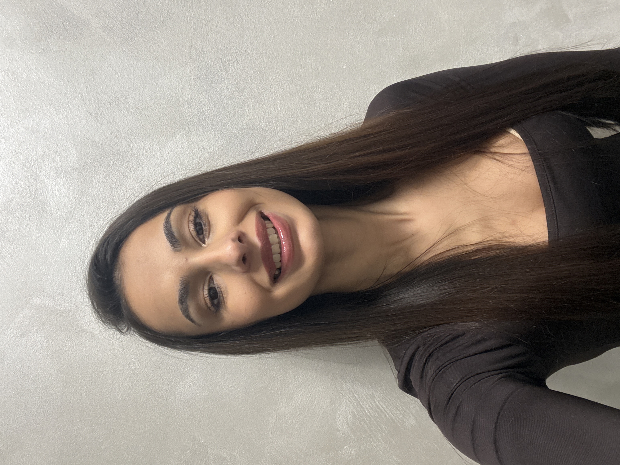 A young woman with long, dark brown hair is smiling at the camera. She is wearing a black, long-sleeved shirt. The background is a light gray wall. There is no indication of the location being Sofia, Bulgaria.