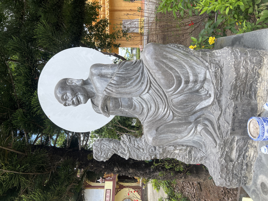 This outdoor image captures a large, grey stone statue within a temple complex in Đăk Mar, Vietnam. The central subject is a statue depicting an elderly, bald male figure with a wrinkled face, prominent ears, and a smiling expression. The figure is seated cross-legged, draped in flowing robes, with its hands in a gesturing posture. A large, plain white circular halo is positioned directly behind the figure's head. The statue rests on a dark, rough-hewn stone base. To the immediate left of the main statue, a smaller, vertically oriented grey stone carving features intricate, swirling patterns, possibly representing a stylized dragon or elemental motifs.
In the foreground, directly in front of the statue's base, a blue and white patterned porcelain incense burner containing dark ash is placed on a paved surface, flanked by two small white ceramic objects.
The background on the left features the trunk and dense green needles of a large evergreen tree. Beyond the tree, a multi-story building with traditional Vietnamese architecture is visible, characterized by yellow, red, and white elements, ornate curved rooflines, and a red decorative lantern.
The background on the right displays a large, golden-yellow temple building with tiered roofs and detailed architectural features. Scattered green foliage and shrubbery, including some with small yellow and red flowers, are present in the mid-ground. A smaller, lighter grey statue is partially visible in the distance to the right of the main subject. The scene is illuminated by clear daylight.