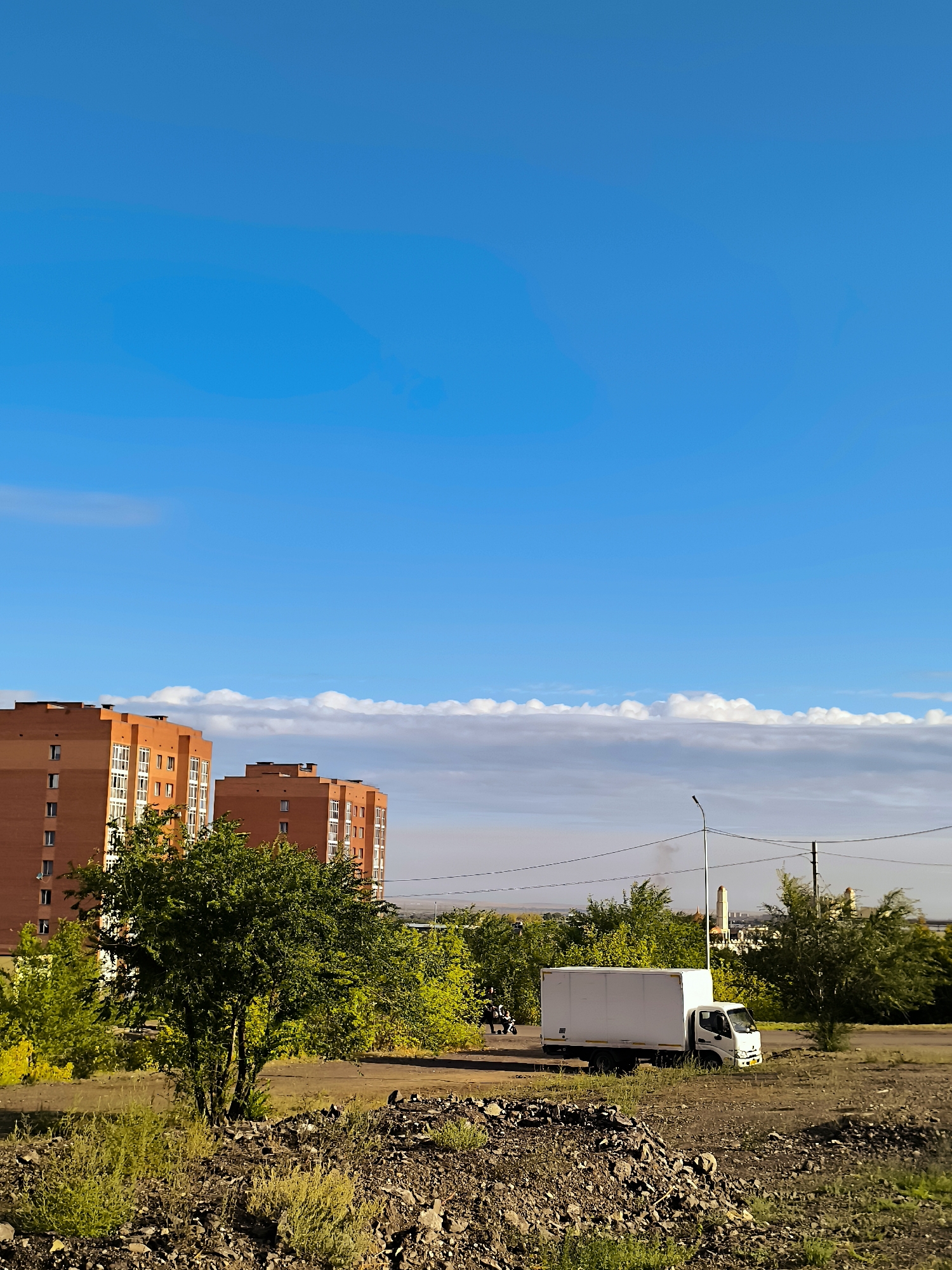 The image shows a residential area in Qarağandı city administration, Kazakhstan. In the background, two multi-story brick buildings are visible, characterized by their orange facades and white-framed windows. In the middle ground, a white box truck is parked on a dirt road. Adjacent to the truck, a single street light stands, and power lines stretch across the scene. Several trees with green foliage are scattered throughout the area, some appearing more densely packed than others. The foreground consists of a rough, rocky terrain with sparse vegetation. The sky above is a clear blue, with a band of white clouds stretching horizontally across the horizon.