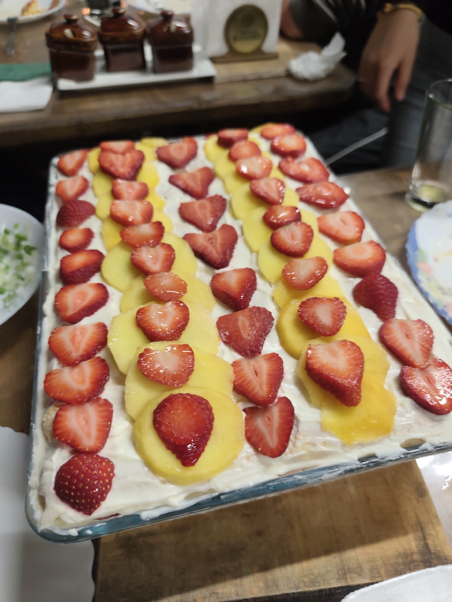 The image depicts a rectangular dessert in a glass baking dish, topped with rows of halved strawberries and circular slices of a yellow fruit, likely mango or peaches, arranged on a creamy white base. The dessert sits on a wooden surface, possibly a table or countertop. In the background, there are small brown ramekins arranged on a white serving platter, and a partial view of a person's arm and torso is visible on the upper right. A plate with chopped green ingredients is visible on the left side of the dessert. A glass of clear liquid and another plate are partially visible in the background on the right side. The overall scene suggests a home or restaurant setting where a dessert is being served or prepared. There is no indication that this image was taken in the city of Mosomishte, Bulgaria.