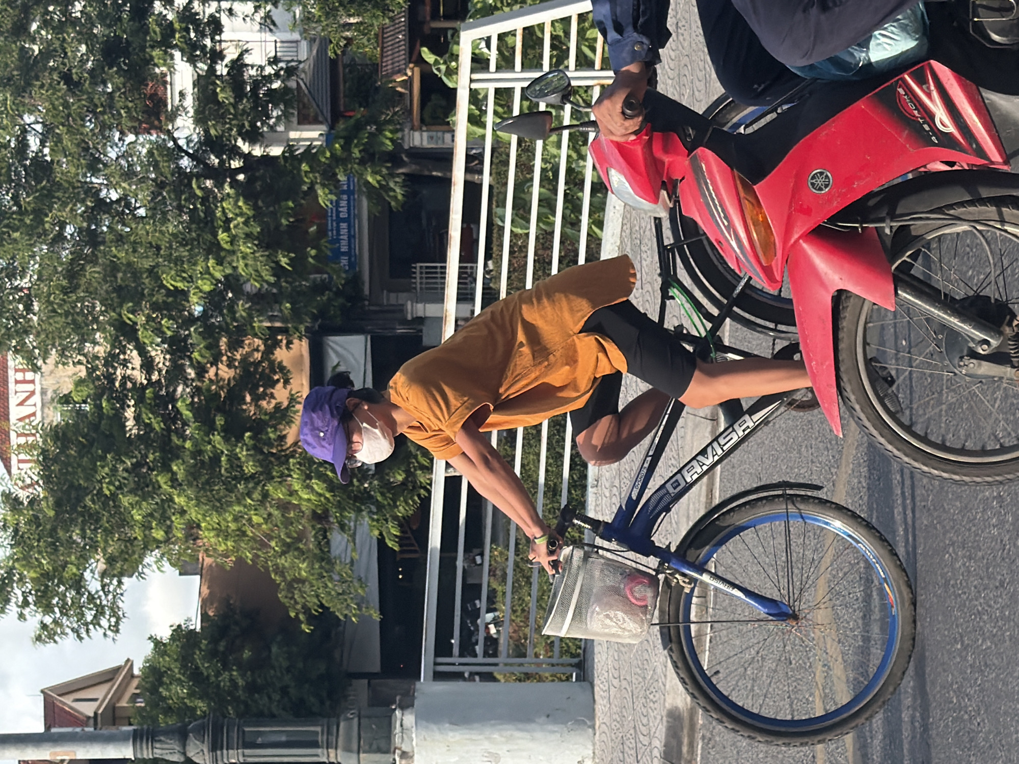 A person wearing a purple hat, glasses, and a white face mask rides a blue bicycle with a white basket in the front. The person is wearing an orange short-sleeved shirt and black shorts. The bicycle has a blue rim on the front wheel. To the right of the bicycle, a red and black motorbike is partially visible. The motorbike is ridden by a person wearing a blue long-sleeved shirt. The scene takes place on a paved road with a white railing in the background, beyond which are trees and buildings. The sky is overcast. The setting appears to be Thuận Hóa Ward, Vietnam, judging by the architectural style and vegetation.