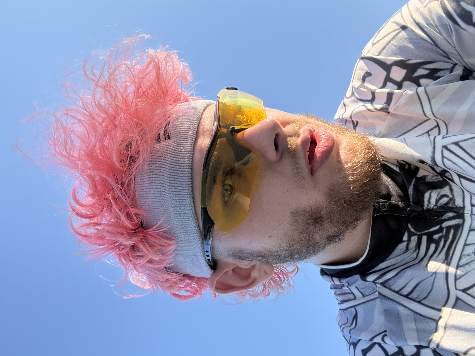 A man with pink hair is wearing a grey headband and yellow tinted glasses. He has a beard and is wearing a patterned jacket with a black collar. The background is a clear blue sky. The image is shot from a low angle looking up. It is not possible to determine if the location is in Đà Nẵng, Vietnam based on the image alone.