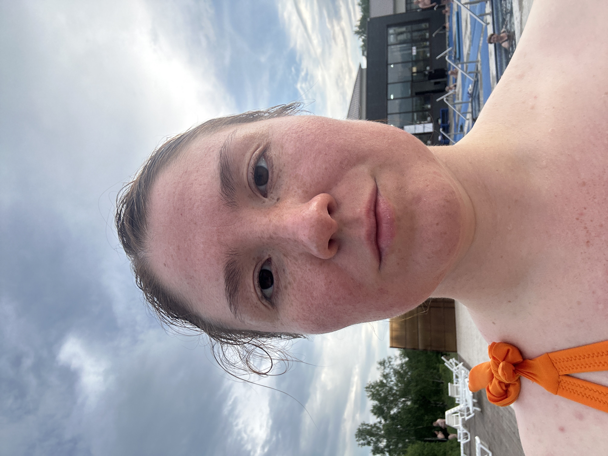 A young woman, with fair skin showing redness and freckles, poses for a selfie outdoors. Her hair is dark and appears damp, with strands falling across her forehead. She is wearing an orange bikini top, adorned with a decorative knot on the strap. The background suggests a recreational setting, possibly a water park or spa, with swimming pools visible and lounge chairs arranged on a wooden deck. In the distance, a modern building with large windows and trees line the perimeter. The sky is overcast with dramatic clouds. The woman's facial expression is neutral.