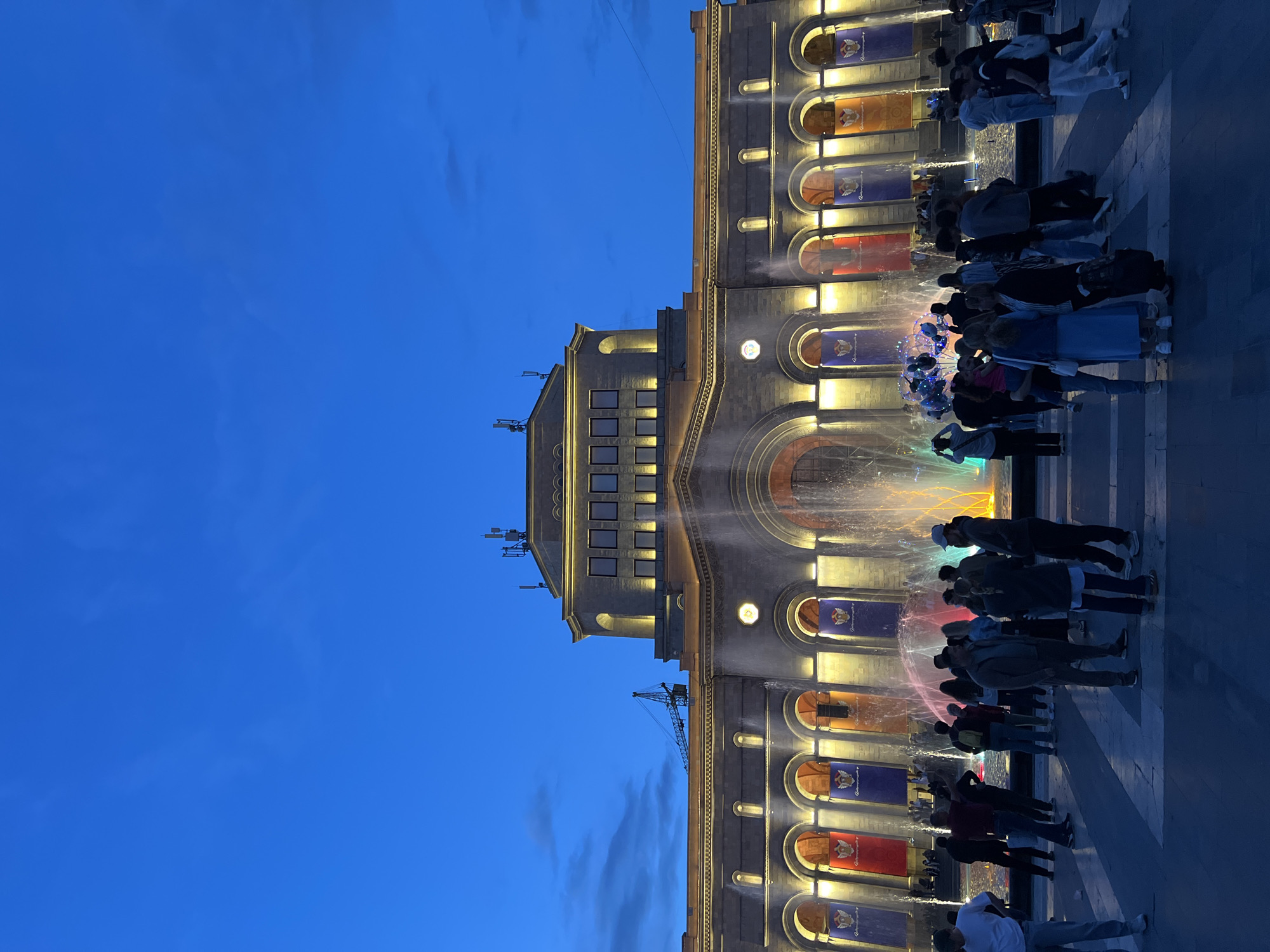 The image shows a building with an illuminated fountain in Yerevan, Armenia, at dusk or twilight. The building is tall, constructed of light-colored stone, and features several arches and windows. Each arch contains a banner. The building's architecture is illuminated with golden light. A water fountain is positioned in front of the building. The fountain is illuminated and consists of multiple streams of water that are various colors. A crowd of people are gathered around the fountain. The sky is a deep blue color. A crane is visible to the left of the building.