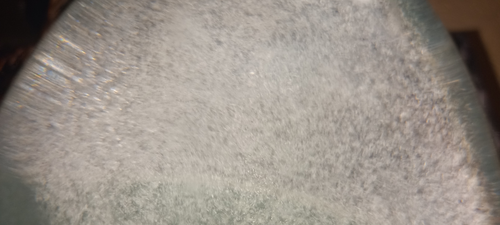 The image shows a close-up, macro view of numerous small bubbles or effervescence within a clear liquid. The liquid appears to be in a transparent container, possibly glass, as suggested by a curved edge and reflections. The lighting causes some prismatic effects and highlights on the bubbles, creating a shimmering and somewhat blurred appearance. The background is dark and out of focus. There are no people or specific identifiable objects or locations visible in the image, making it impossible to determine its context within Kryvyi Rih, Ukraine. The scene is purely abstract, focusing on the texture and optical properties of the bubbly liquid.