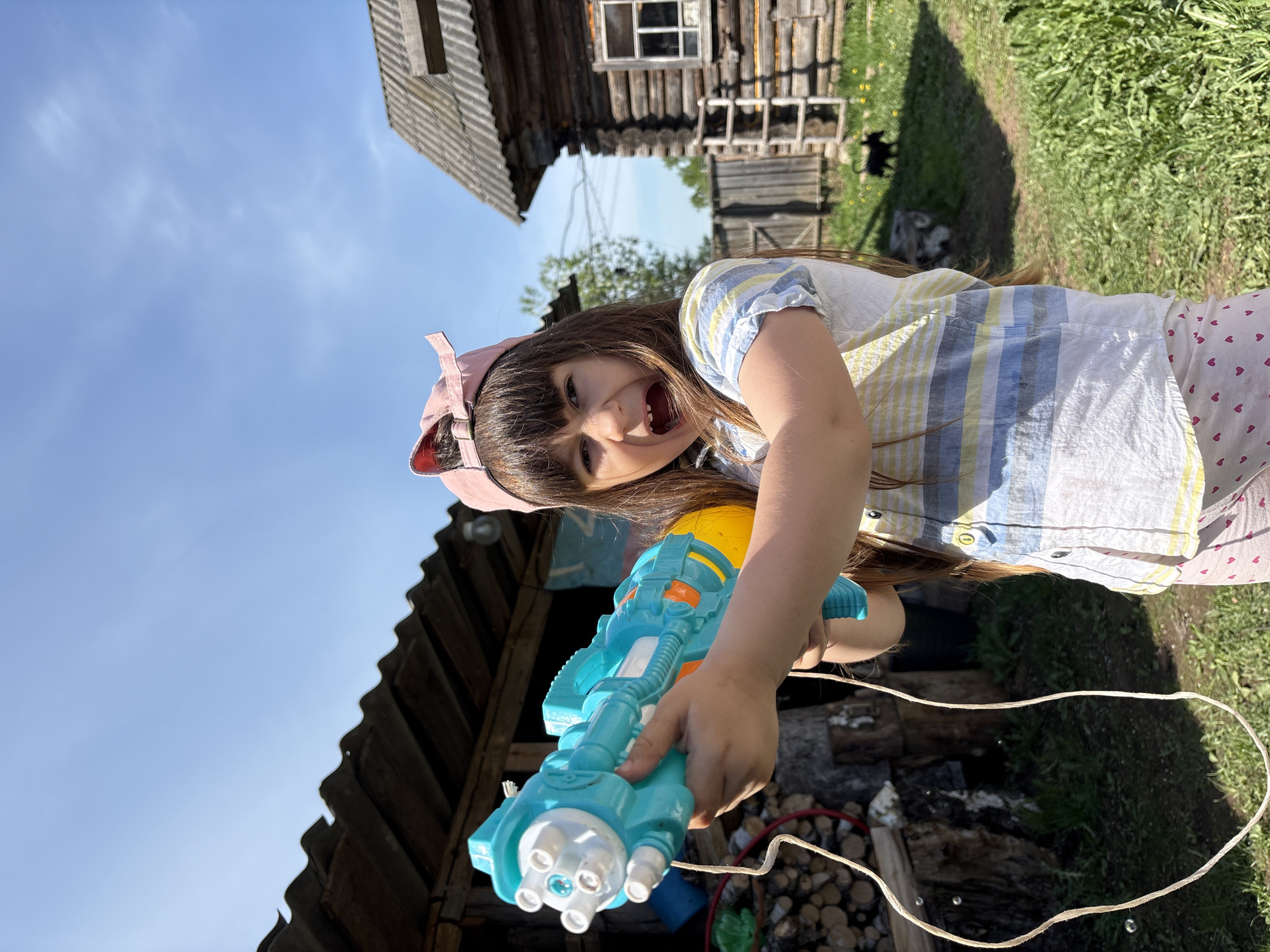 A young girl, possibly in Nizhnyaya Chermoda, Russia, aims a blue and yellow toy gun at the camera. She wears a pink cap and a striped shirt, and is standing in a yard with wooden buildings in the background. Firewood is visible near the girl. The sky is clear.