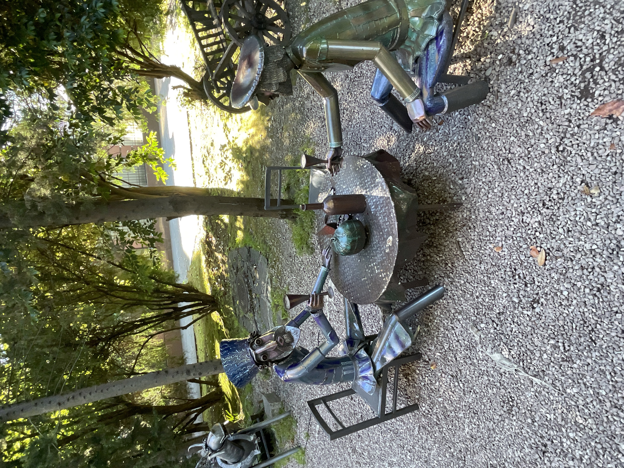 This image shows two metal sculptures of men sitting at a round table in an outdoor setting. The sculptures are dressed in period clothing, one in blue and the other in green, with distinctive hats. Both are holding glasses, and there are bottles and a jug on the table. The sculptures are sitting on chairs at a metal table covered with small stones. The background includes trees, gravel, and a hint of a paved area and a wooden cart. The lighting suggests it is daytime. The location is the Arboretum Botanical Garden in Sochi, Russia.
