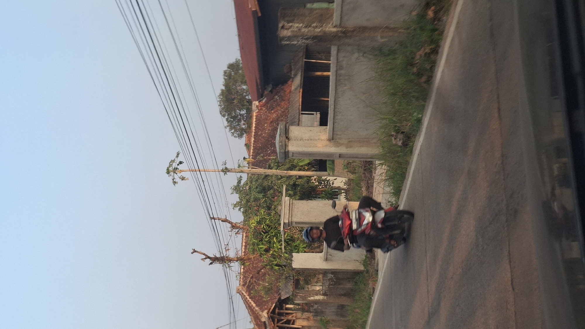 A person wearing a black shirt and a blue helmet rides a red and white motorcycle on a paved road. The motorcycle has a silver front fender and silver accents on the body. The rider is looking forward. The road has some shadow patterns on it. To the left of the road, there is a concrete fence with a gate. Behind the fence, there are houses with terracotta tile roofs and some trees. One house has a wooden structure in front of it. To the right of the road, there is a concrete wall with posts and some overgrown grass. In the background, there are power lines stretched across a pale blue sky. A tall, thin tree with sparse leaves is visible behind the power lines. The image appears to be taken in Cibinong Hilir, Indonesia, judging by the architectural style of the houses and the presence of power lines.