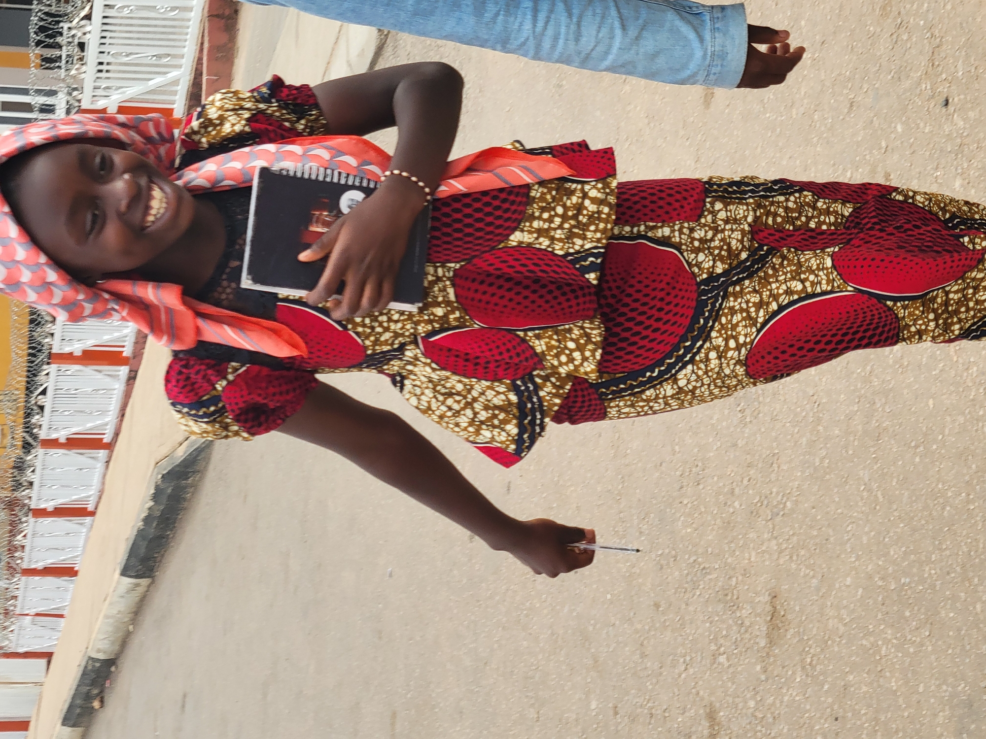 A young woman smiles while standing on an asphalt surface, holding a pen in her right hand and a closed notebook in her left. She is wearing a patterned dress of red, brown, and black with a matching headscarf. A bracelet adorns her left wrist. Above her, out of focus, is a person wearing jeans. A white fence with orange trim runs along the left edge of the frame. The location is suspected to be Jos, Nigeria.