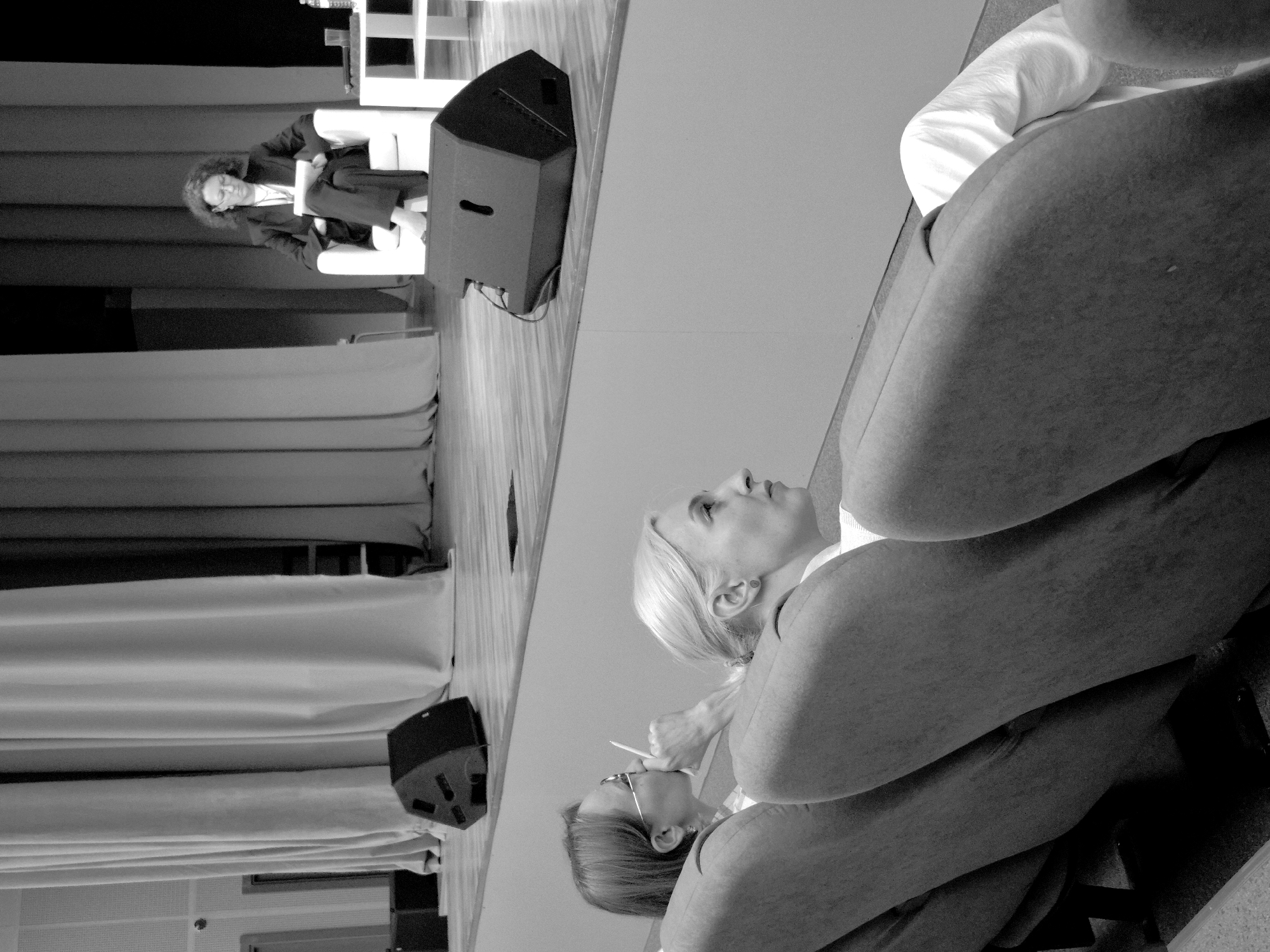 The black and white photograph, taken in Kungur, Russia, depicts a conference or presentation setting. Two women are the primary subjects. One woman, with curly hair and glasses, is seated in a chair on the left side of the frame, looking forward. She is wearing a dark jacket and pants. In the foreground, another woman with blonde hair is seen from the side, looking up and to the right. She is wearing a white top and a dark jacket. A third person's arm and shoulder, wearing a white sleeve, is visible in the upper right of the frame.
The scene includes a stage area with a podium or lectern on the left, and a large speaker is visible. Drapes or curtains hang in the background, creating a formal atmosphere. The lighting suggests an indoor event, possibly during the day. The expressions of the subjects appear attentive and engaged with the proceedings. There is no visible text in the image.