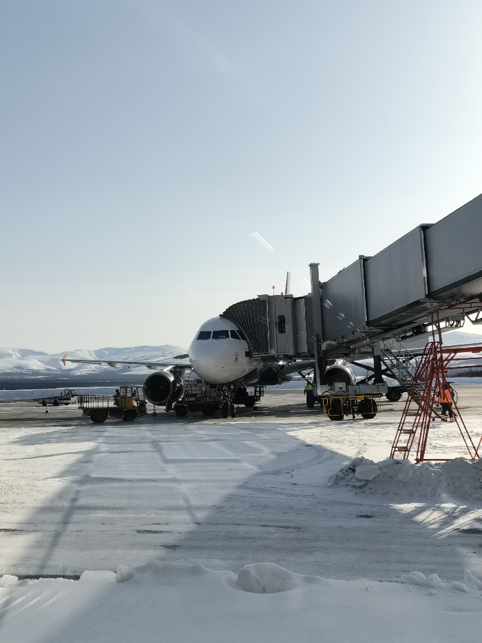 A white narrow-body passenger jet, likely an Airbus A320 series aircraft, is parked on a snow-covered tarmac at Sokol Airport, near Magadan, Russia, under a clear, bright blue sky. The aircraft's forward port side is connected to a grey, accordion-style jet bridge extending from an unseen terminal building to the right.
Ground support equipment is positioned around the aircraft: a yellow ground power unit or baggage tractor with a trailer is located under the nose, and a yellow cargo loader is positioned under the forward fuselage. A third yellow ground vehicle is further to the left of the aircraft. A red multi-level maintenance stand or ladder is visible in the foreground on the right, with one individual in an orange high-visibility jacket standing on it. Another individual in a yellow-green high-visibility jacket is near the jet bridge attachment point to the aircraft.
The tarmac is partially cleared but predominantly covered in patches of snow and ice, with visible tire tracks. Significant snowdrifts are present in the immediate foreground. In the distant background, snow-covered mountains or hills are visible on the horizon. Contrails from a distant aircraft are faintly discernible in the upper portion of the sky. The scene is illuminated by strong sunlight, casting distinct shadows of the jet bridge and aircraft onto the snow.