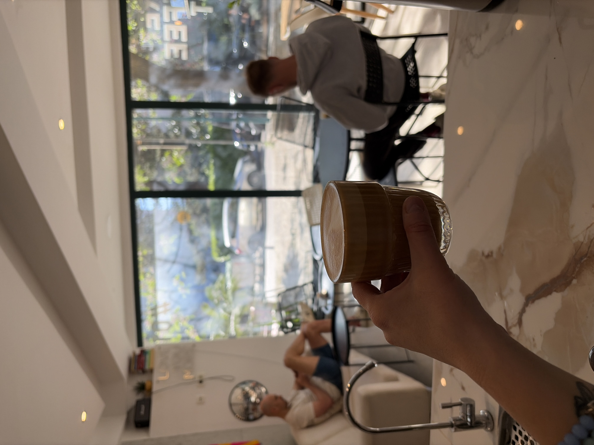 A person's hand holds a glass of coffee. The photo is taken inside a building in Budva, Montenegro. Through a large window, people are visible outdoors in the street, one sitting at a table. In the background, inside, there's another person reclining on a couch. A sink and counter are also visible.