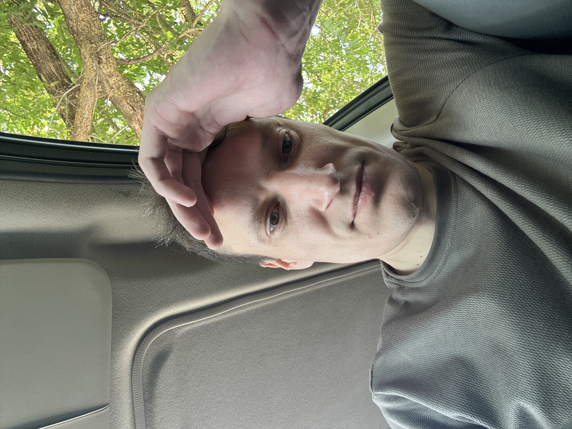 The image shows a close-up, selfie-style shot of a man's face and upper body, taken from inside a vehicle. The man is wearing a grey, textured long-sleeved shirt. His right hand is resting on his forehead, and his gaze is directed downwards. The interior of the vehicle, including a grey sun visor and part of the ceiling, is visible. Through an open window on the right side of the frame, lush green foliage and tree branches are apparent, suggesting an outdoor setting adjacent to the vehicle. The lighting suggests it is daytime. There is no specific indication of the location being in Makiv, Ukraine.