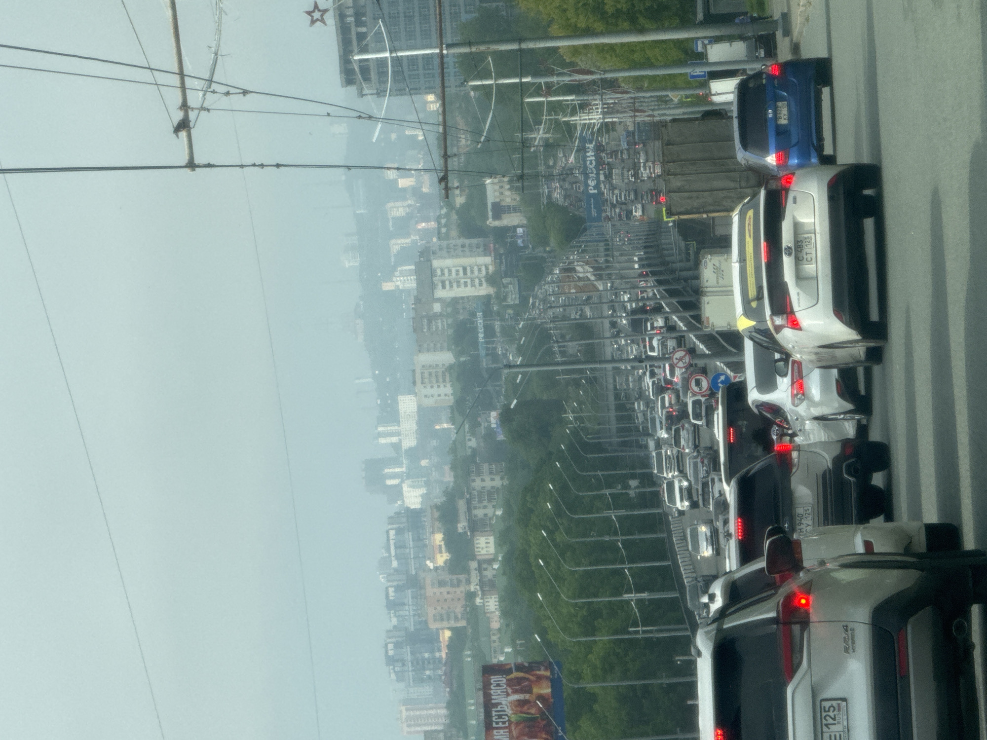 The image depicts a congested urban highway during daytime, likely in a city with a hilly terrain and a distant skyline. The foreground features a traffic jam with numerous cars in multiple lanes. The vehicles are of various makes and models, with some prominent ones including a white Toyota RAV4, a white Toyota Prius with a yellow roof, and a blue Honda Fit. The road appears to be an elevated highway or bridge, indicated by the presence of lampposts and the elevated perspective. In the background, a dense cityscape with numerous buildings of varying heights is visible, shrouded in a hazy atmosphere. There are also several billboards and advertisements along the side of the road. The sky is overcast, contributing to the diffused lighting conditions. Power lines and utility poles are visible overhead, suggesting the presence of electrical infrastructure. The overall scene conveys a sense of urban sprawl and heavy traffic flow.