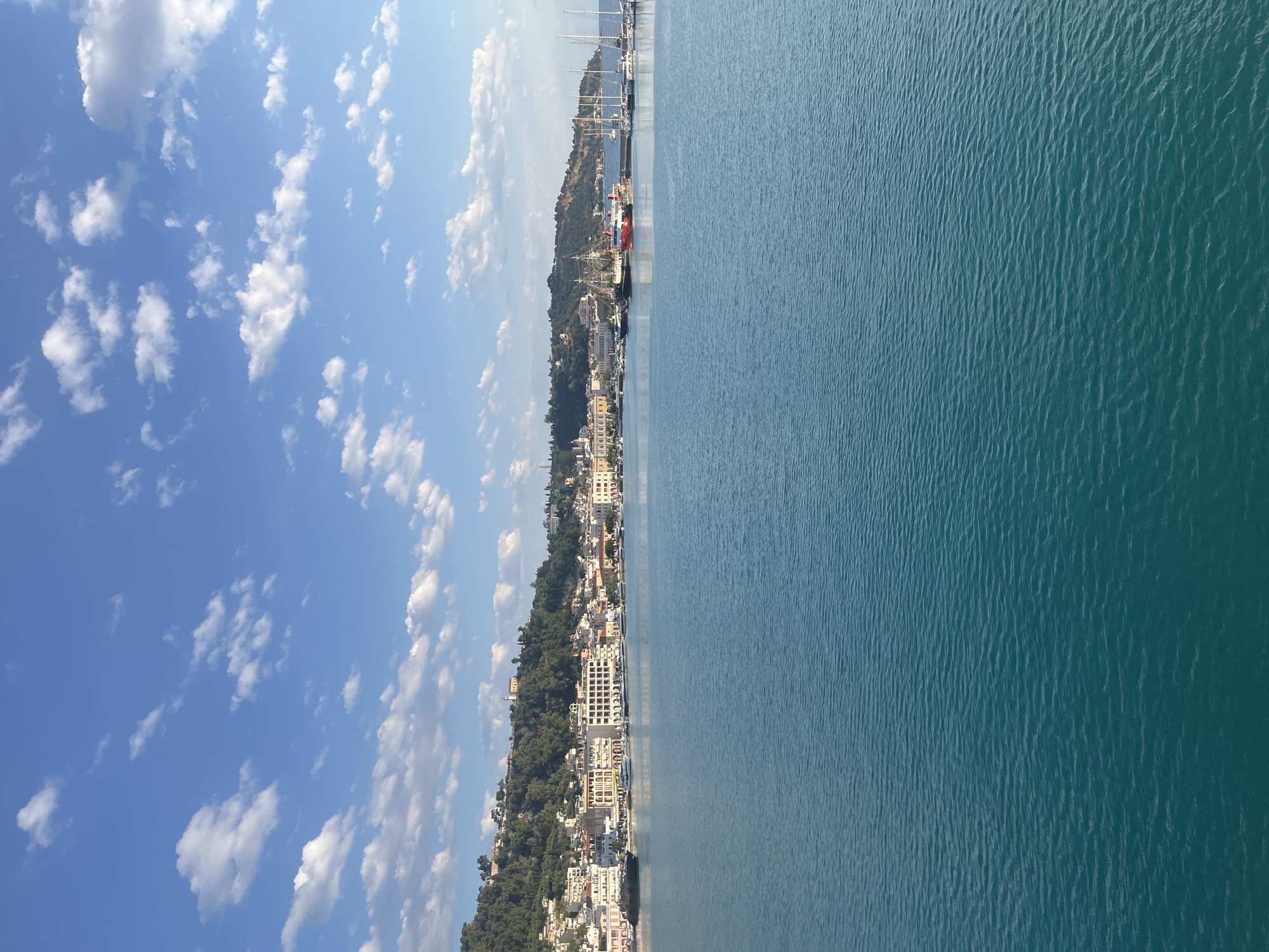 The image depicts a scenic view of the Zakynthos harbor in Zakynthos Municipal Unit, Greece. The foreground is dominated by the clear, turquoise waters of the Ionian Sea, with gentle ripples reflecting the sunlight. In the middle ground, a waterfront promenade is lined with buildings, featuring a mix of architectural styles with predominantly white and terracotta facades. Several boats and yachts are docked along the harbor, including a notable sailing ship with prominent masts and a red and white vessel. The background consists of a lush green hillside covered in trees and dotted with buildings and houses, extending towards the top of the frame. The sky above is a vibrant blue, adorned with scattered white, puffy clouds. The overall scene conveys a peaceful and picturesque coastal setting.