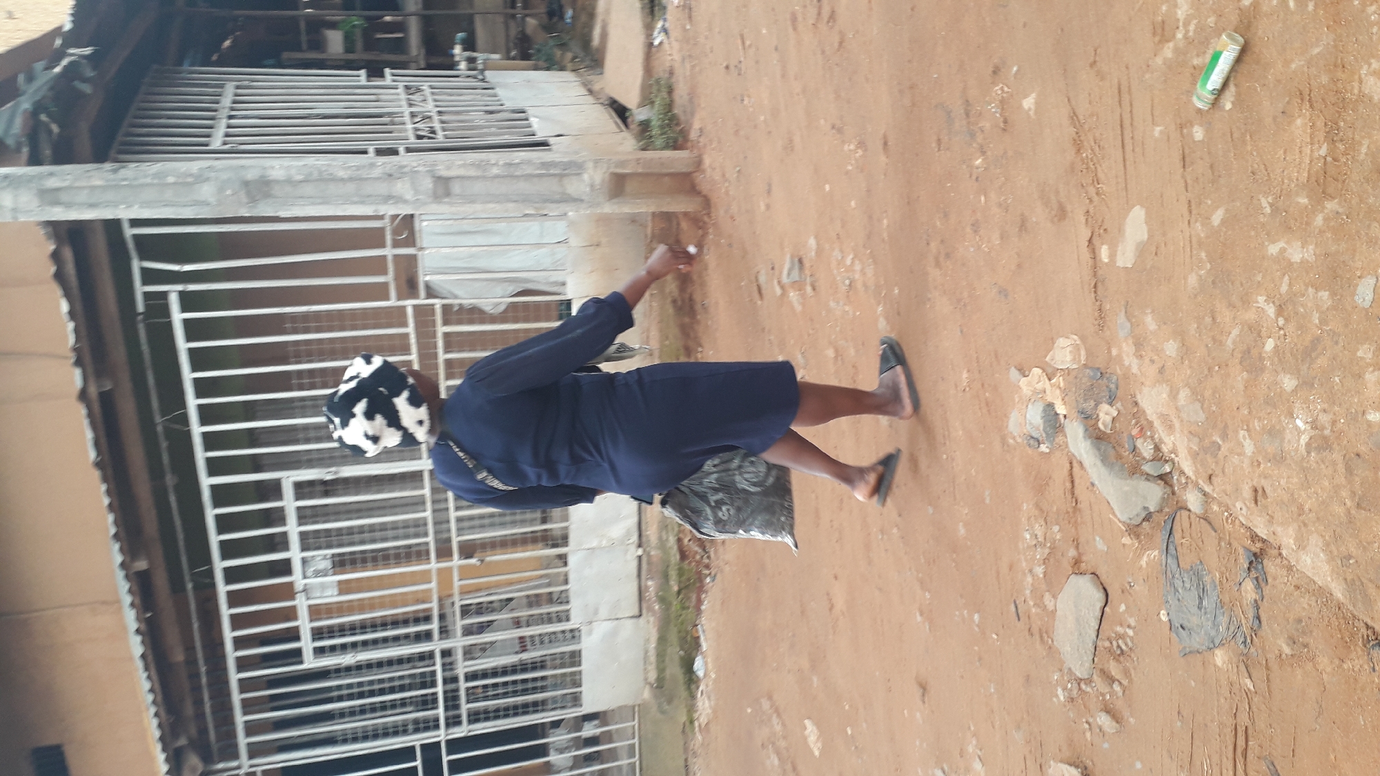 A person walks along a dirt road in Nkpor, Nigeria. The person is wearing a dark blue dress and a black and white patterned hat. They are carrying a dark bag over their left shoulder and a plastic bag in their right hand. The road is unpaved and appears dusty. To the left of the road, there is a building with a metal gate and a corrugated metal roof. A light-colored pole stands to the right of the person. Debris, including rocks and plastic, is scattered on the ground. A green and white bottle lies on the ground to the right. The scene is outdoors, with natural lighting.