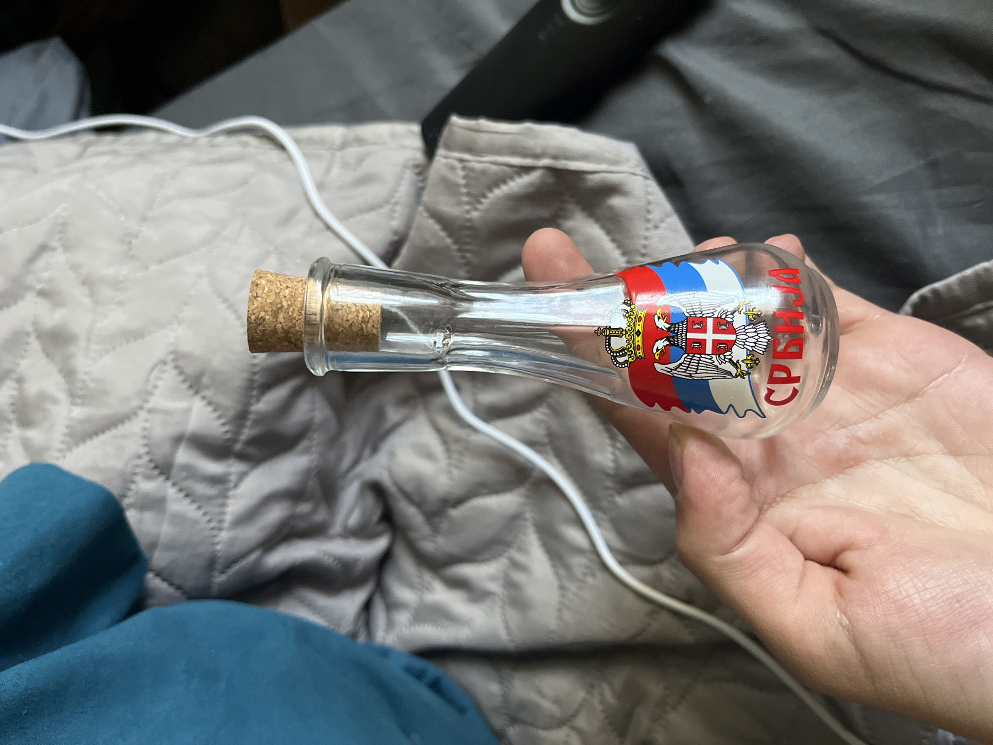 A clear glass bottle with a cork stopper is being held in a person's hand. The bottle has a Serbian coat of arms and the Cyrillic word "СРБИЈА" (Serbia) printed on it. The background is a quilted gray blanket with a white wire and a blue garment visible. The scene's location in Sofia, Bulgaria cannot be determined from the image.