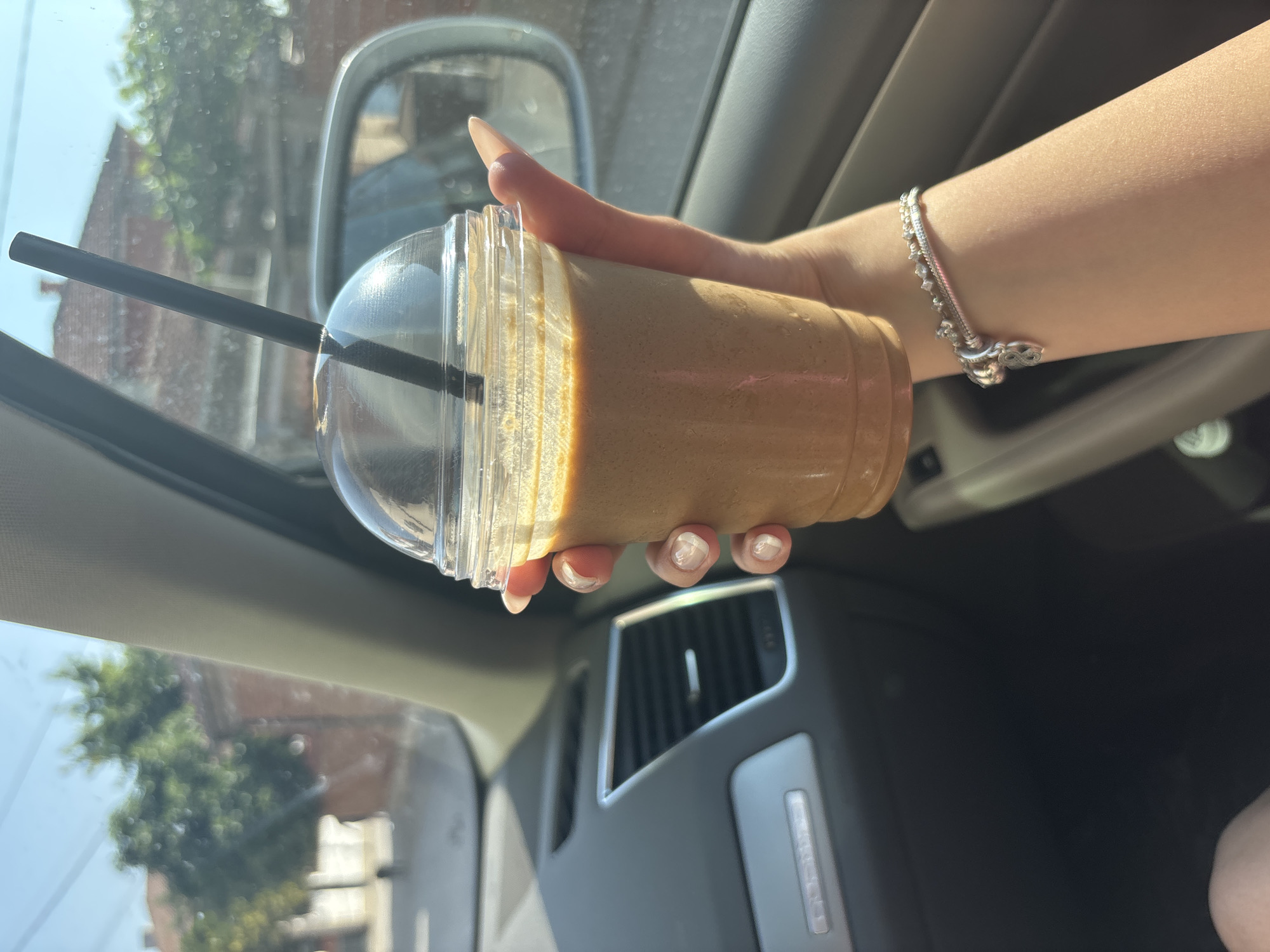 A person's right hand, with manicured fingernails, holds a clear plastic cup containing a light brown, frothy beverage, possibly an iced coffee or smoothie, through which a black plastic straw protrudes from a dome lid. The hand wears a silver-colored charm bracelet with multiple charms, including one resembling an infinity symbol. The scene is set inside a vehicle, likely an Audi Q5 based on visible dashboard elements and an air vent. Sunlight illuminates the interior from the right. The exterior environment, viewed through the passenger-side window and reflected in the side mirror, is a blurred street scene in Rakovski, Bulgaria. It shows residential buildings with reddish-brown tiled roofs, green trees, utility wires, and a clear blue sky, indicating a sunny day.