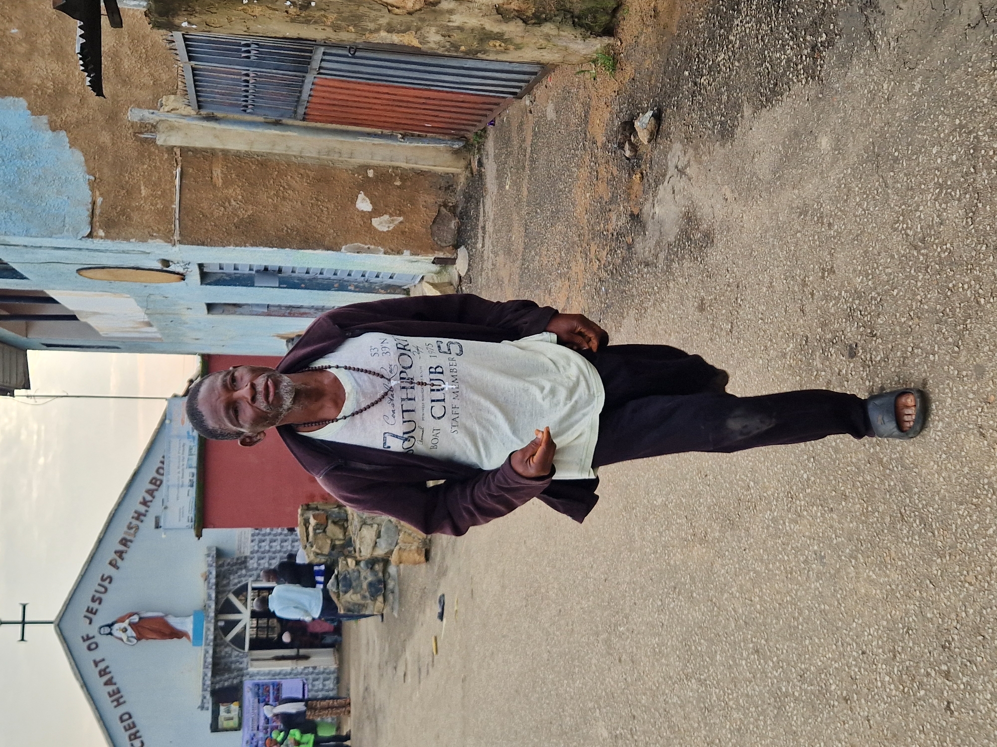 A man walks towards the camera on a paved street in Kabong, Nigeria, with a church building in the background. The man is middle-aged, with dark skin, a graying beard, and is wearing a dark jacket over a white t-shirt and dark pants. He has a beaded necklace and is wearing dark sandals. The church building, identified as "SACRED HEART OF JESUS PARISH KABONG," is light blue with a red lower section and features a statue of Jesus above the entrance. Several other people are visible near the church entrance, some in the distance. The street surface is asphalt with some patches of dirt and scattered stones. Adjacent to the church is a building with a textured brown facade and a metal gate. The sky is overcast.