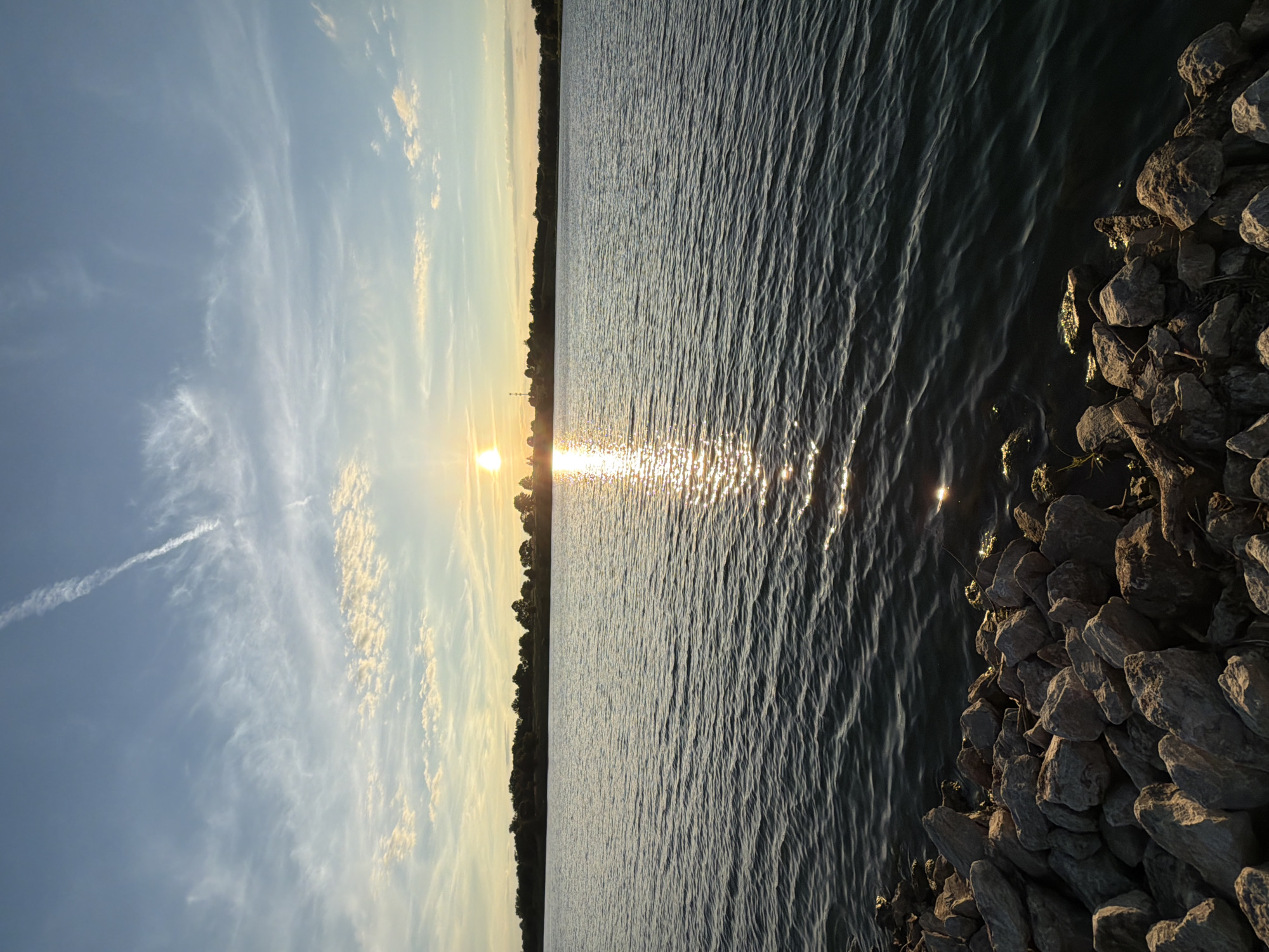 This is a vertical, eye-level, full shot of a body of water with a sunset visible in the background. The foreground is made up of rocks and a bit of grass along the water's edge. The water is dark and choppy, with ripples and the reflection of the sun creating a glittering effect. The sun is setting just above the tree line of a distant shore, casting a warm glow across the sky and water. Wispy clouds dot the blue sky, with a contrail visible high above. The overall scene depicts a serene sunset over a lake or large body of water.