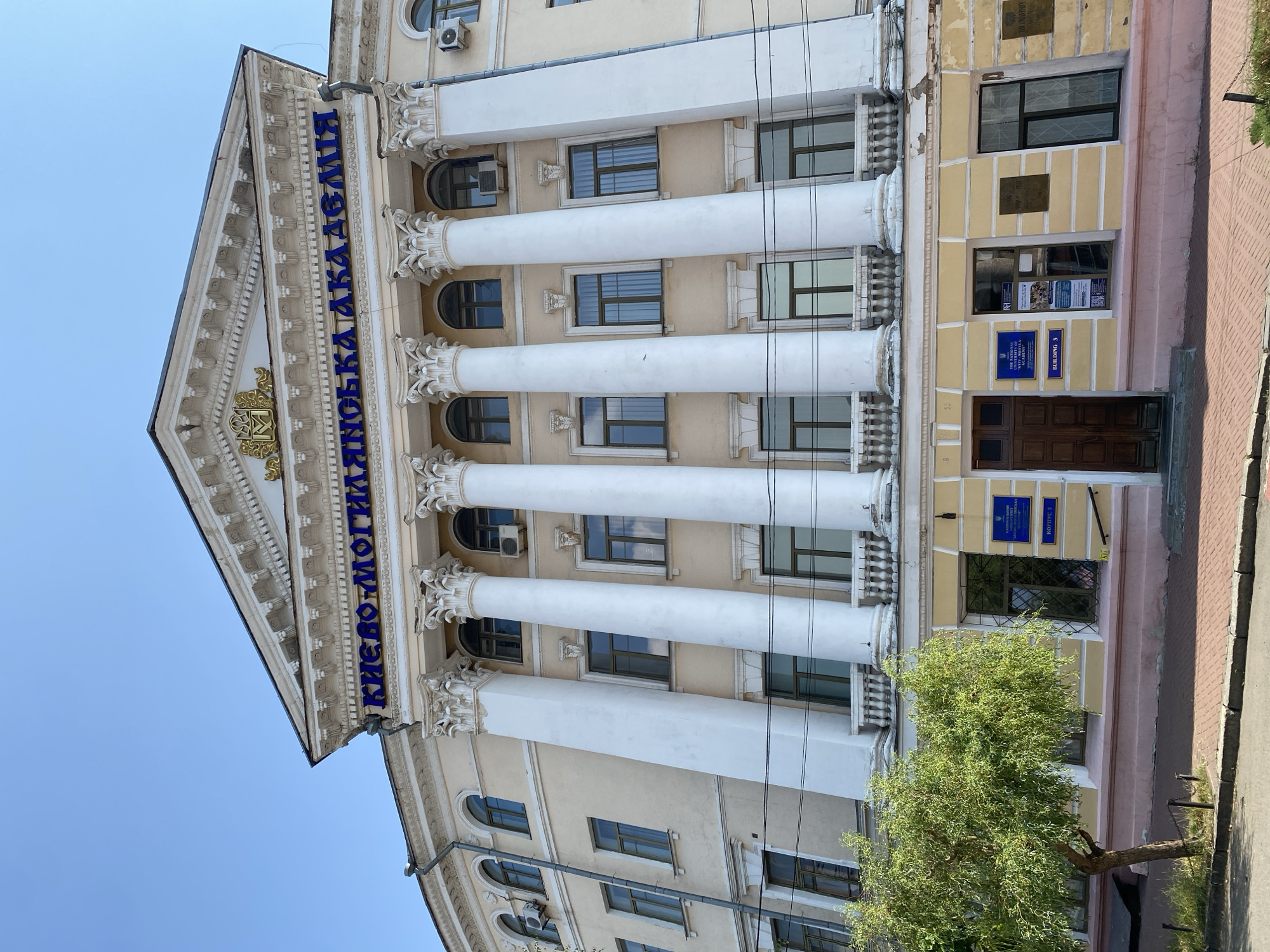 The image shows the facade of a large, classical-style building under a clear blue sky. The building is painted a pale yellow with white Corinthian columns supporting a pediment. Above the columns, blue lettering reads "КИЄВО-МОГИЛЯНСЬКА АКАДЕМІЯ" (Kyiv-Mohyla Academy). Below this, a golden emblem is visible within the pediment. The lower part of the building has a light pink base and several windows. To the left of the building, a small tree with green foliage stands on a paved sidewalk. The scene appears to be a sunny day in a city, likely an academic institution given the lettering.
