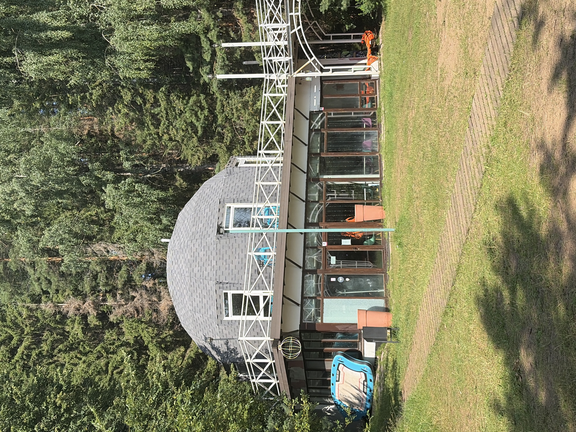 The image shows a unique geodesic dome building with a large glass-walled lower level, situated in a wooded area. The building has a gray shingled dome roof with several windows. A white railing encircles a balcony on the dome level. The lower level features numerous large windows, some of which are reflective. Outside the building, to the left, is a large blue inflatable object. To the right, a white arched walkway connects to the upper level. The area is covered in grass, with a pathway made of wooden planks leading towards the building. Tall green trees surround the structure, suggesting a natural or park-like setting. No people are visible in the image. The location is identified as Kostovaty, Russia.